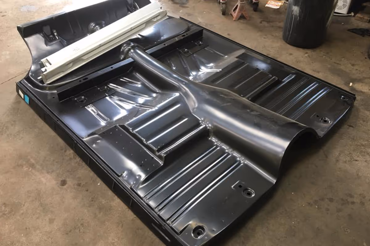 A shiny, black car floor pan lies on a garage floor, showing its various ridges and contours.