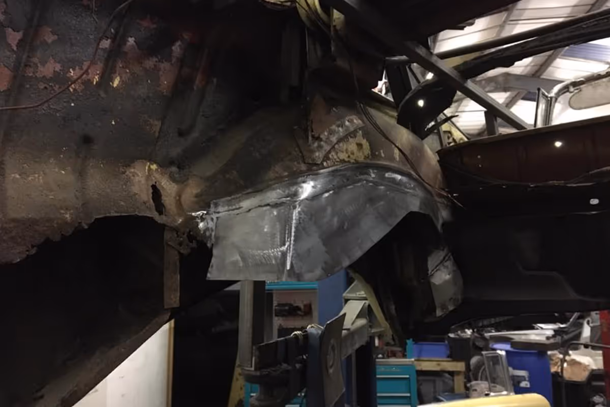 A rusted car frame is undergoing repair, with a newly welded metal patch attached.
