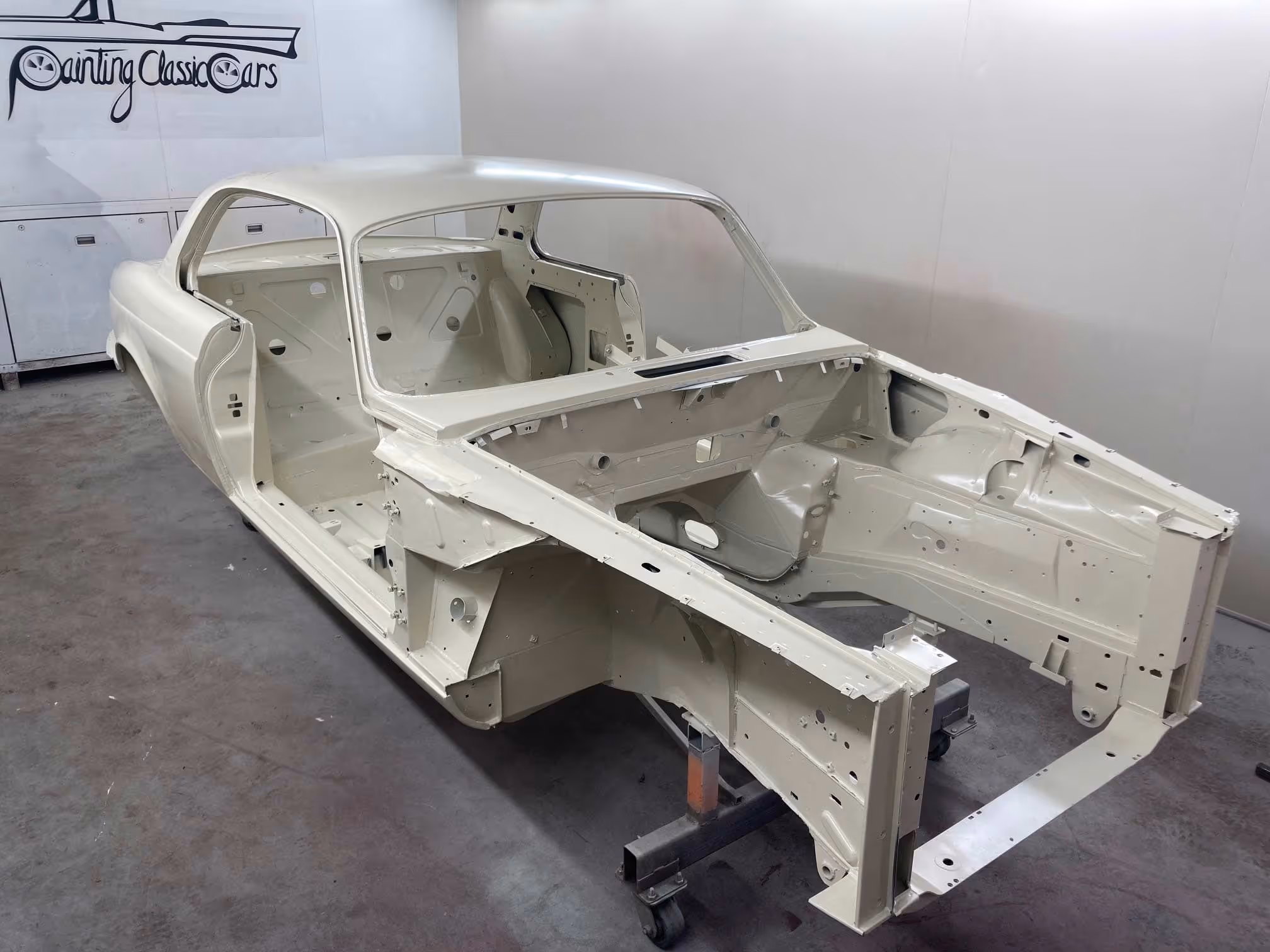 A partially assembled car frame sits in a garage, elevated on a metal stand.