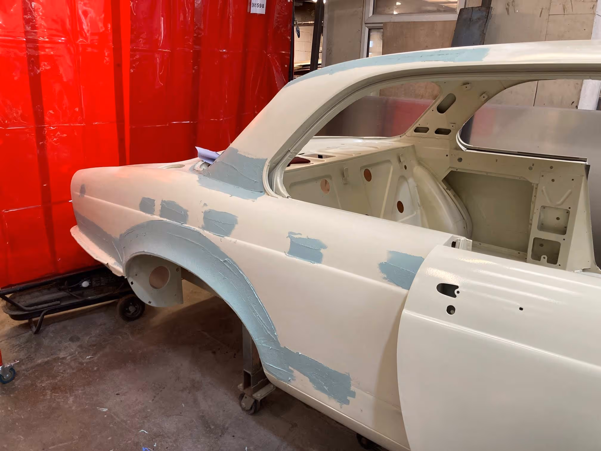 A partially restored car with primer patches is in a workshop near red curtains.