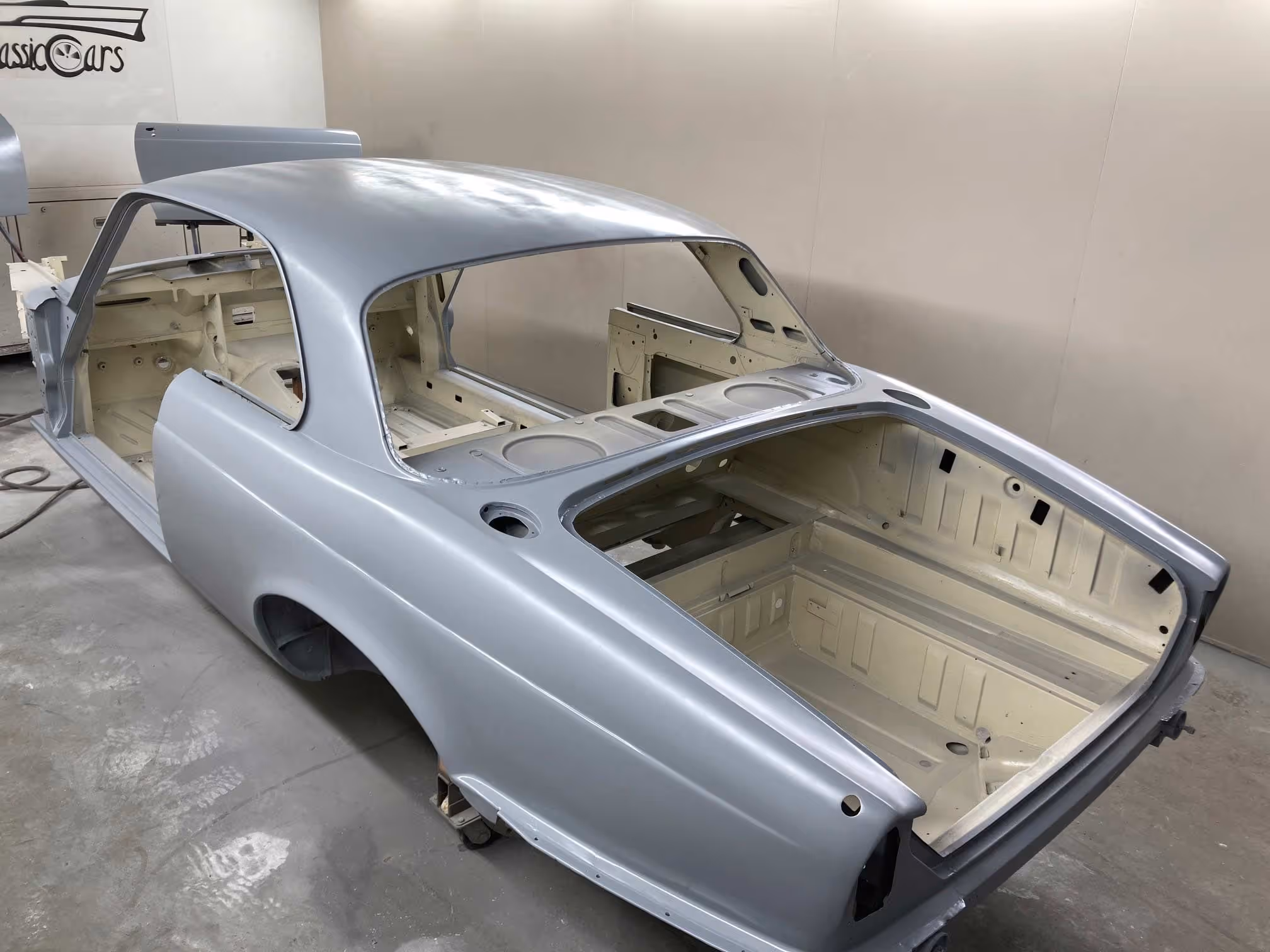 A silver classic car with no interior or doors is being restored in a garage.