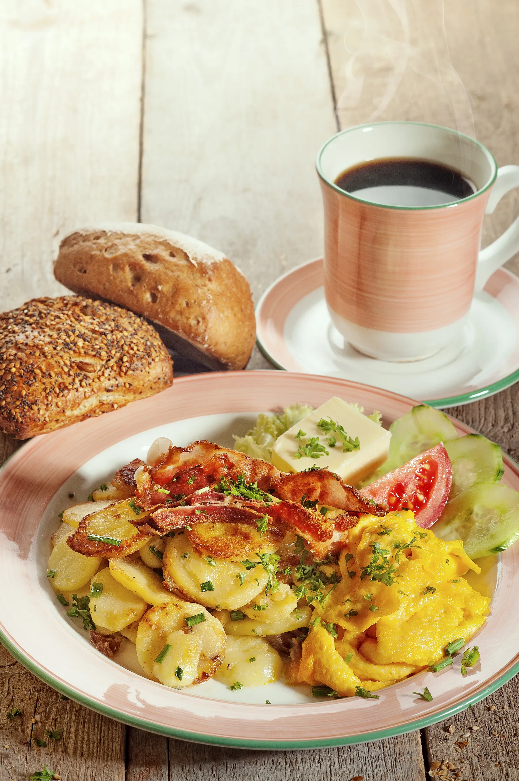 Ein herzhaftes Frühstück mit Kartoffeln, Speck, Tomaten und Gurken. Daneben zwei Brötchen und ein Kaffee.
