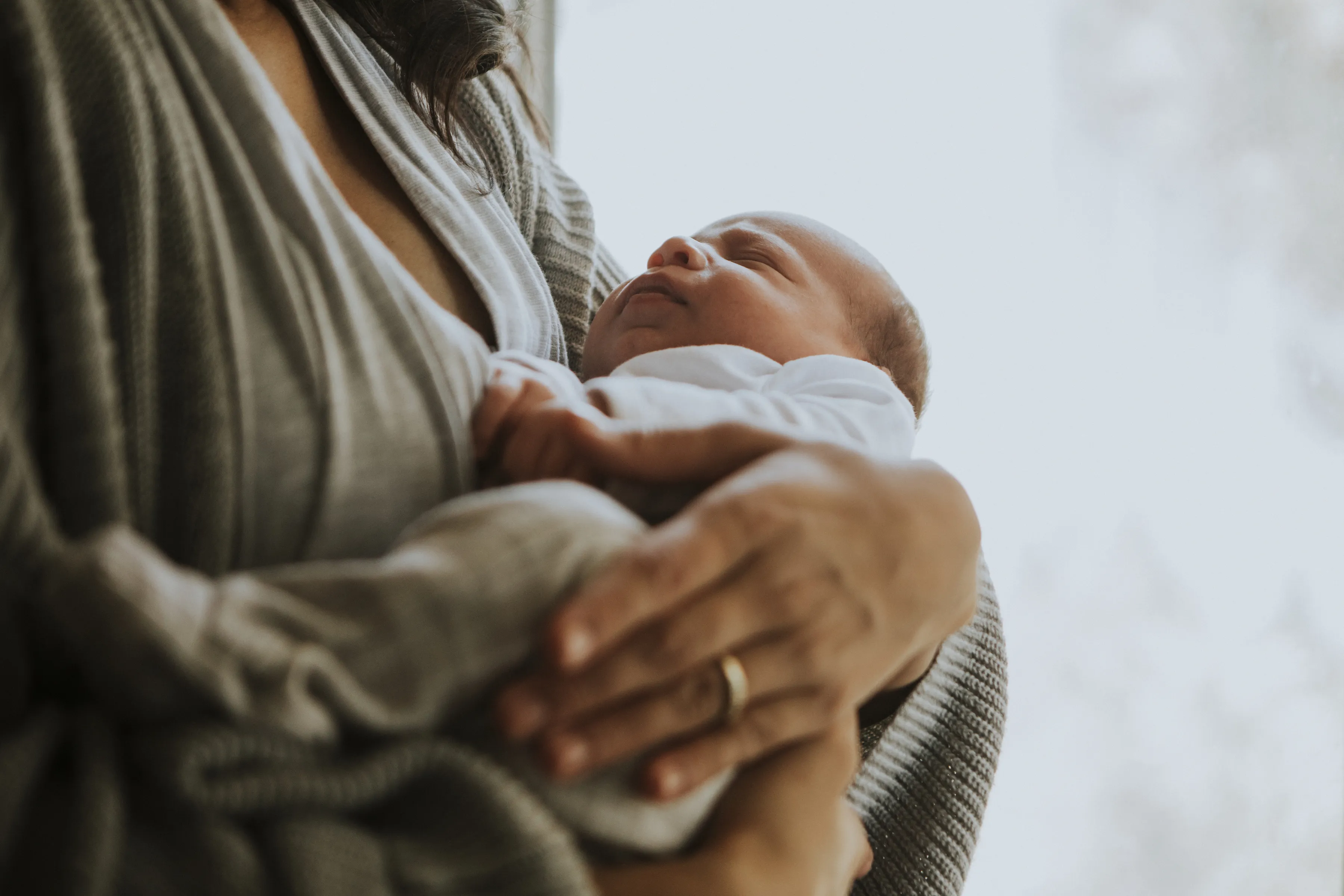 Close-up of a person gently cradling a sleeping newborn baby wrapped in a white outfit.