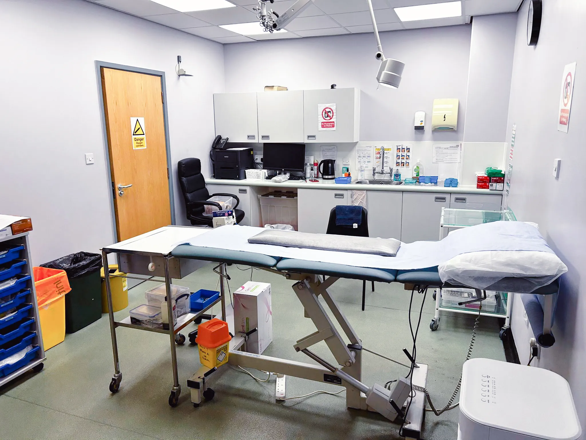 Clean medical examination room with adjustable examination table, office chair, medical supplies, and sink.