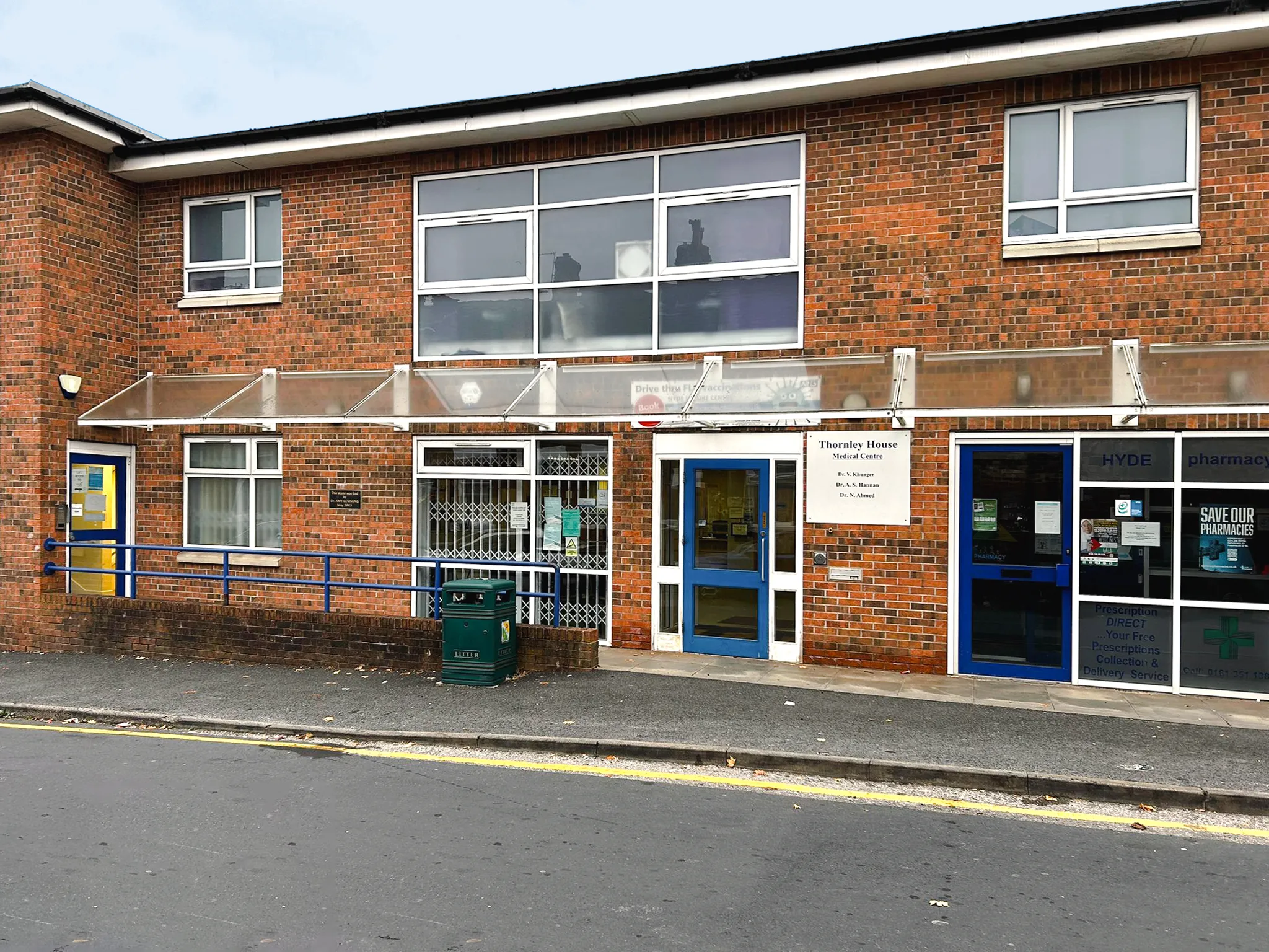 Brick building with blue-trimmed doors housing Thornley House Medical Centre and a pharmacy, with a green litter bin on the sidewalk.