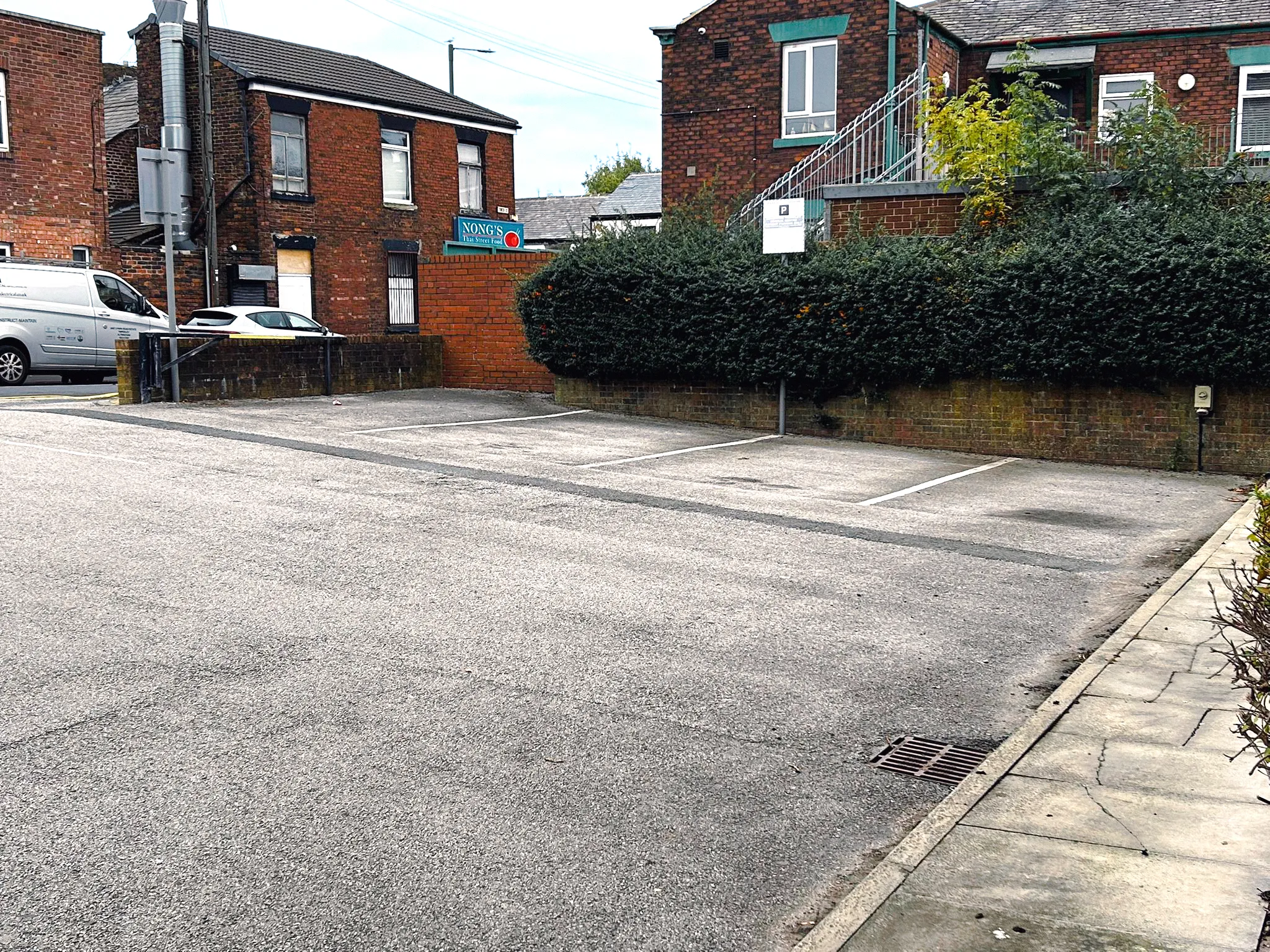 Empty paved parking lot with marked spaces next to brick buildings and dense shrubbery.