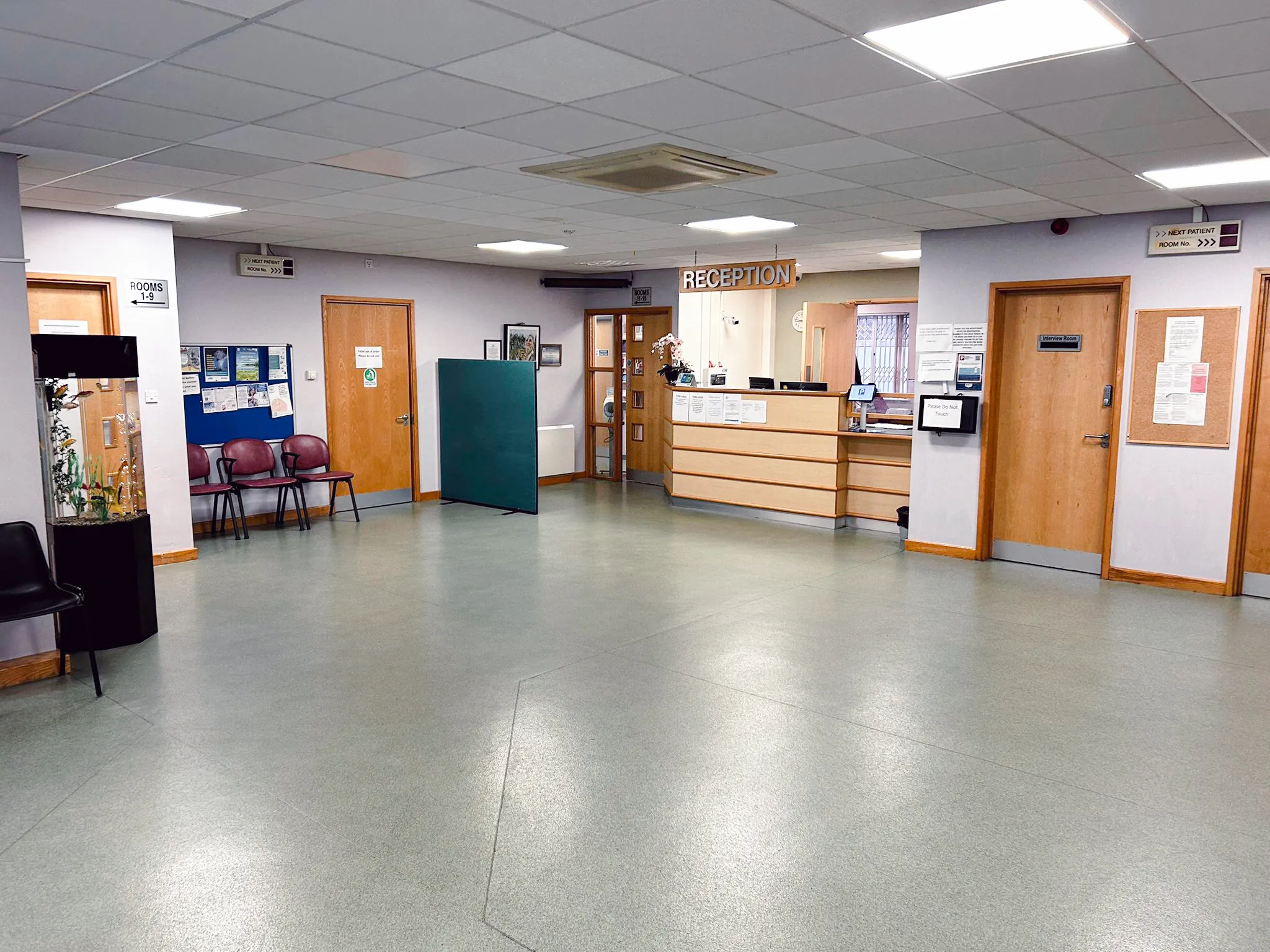 Spacious medical reception area with wooden reception desk, multiple closed doors, green partition, chairs, and informational signs on walls.