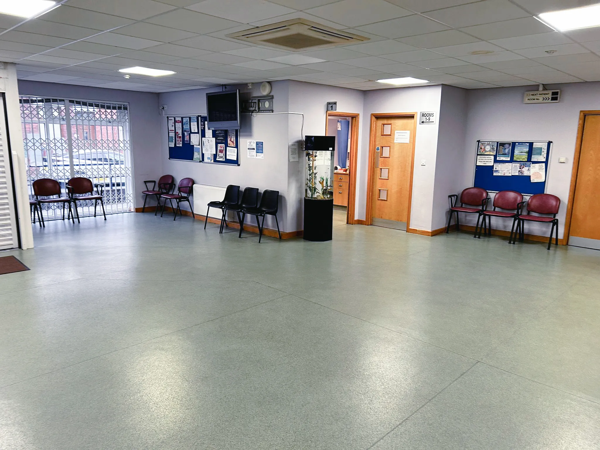 Empty medical clinic waiting room with multiple chairs, bulletin boards, a fish tank, and doors labeled for rooms.