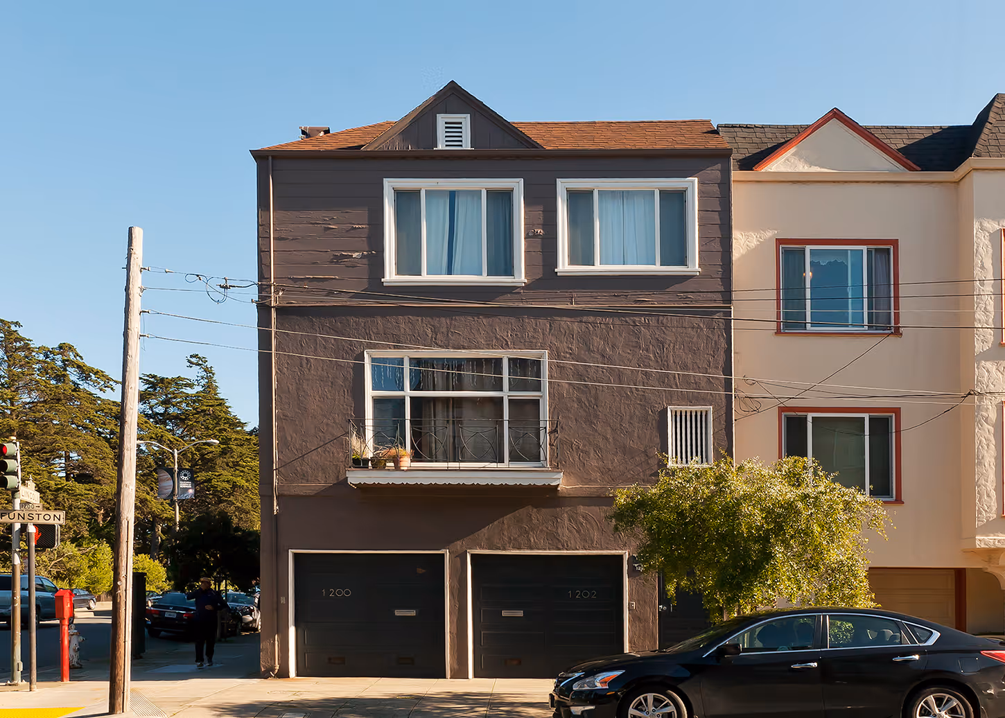 Three-story brown residential building with two garage doors numbered 1200 and 1202, two upper floor windows, a small balcony with plants, and a black car parked in front.