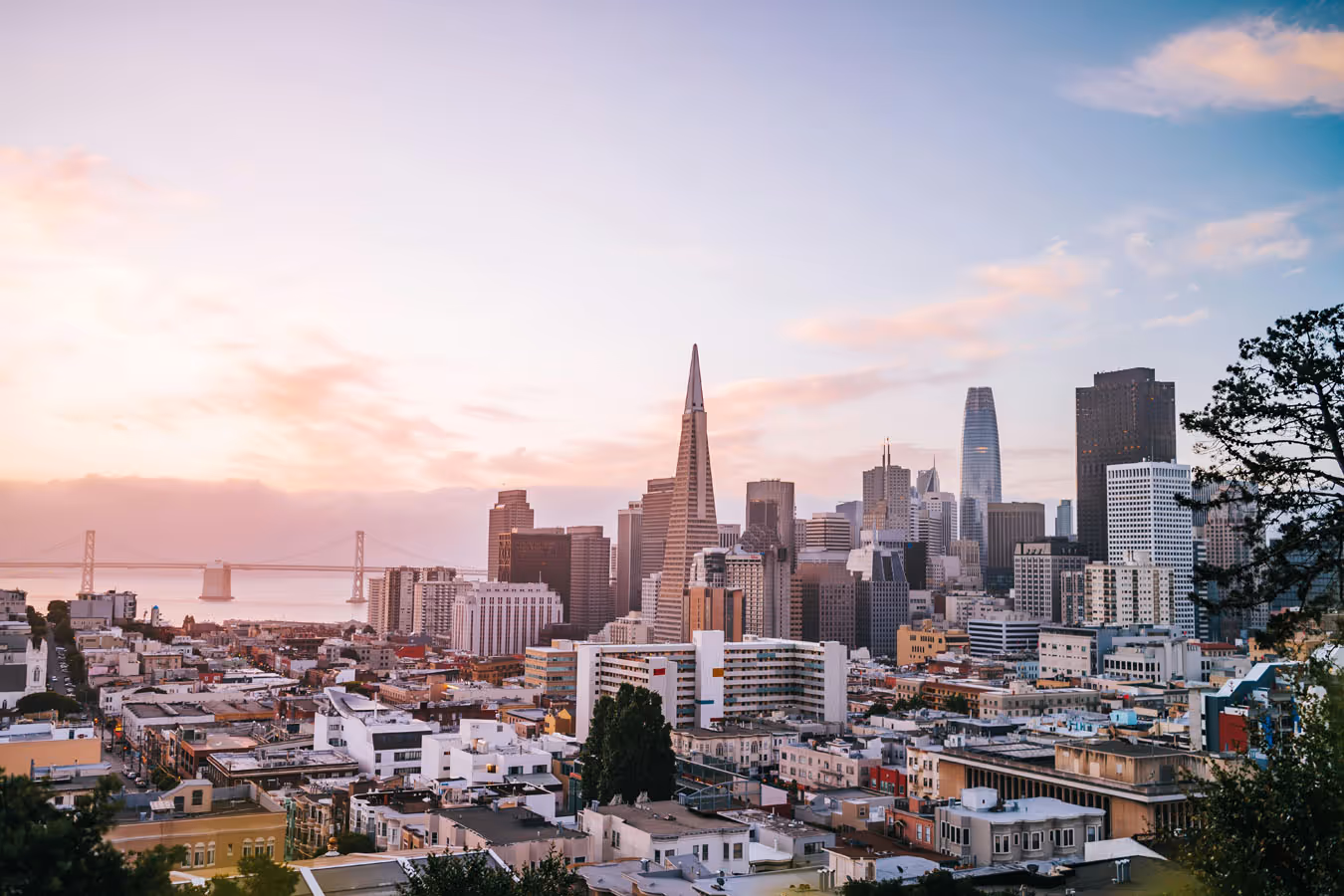 San Francisco skyline at sunset with the Transamerica Pyramid and Bay Bridge under a pink and blue sky.