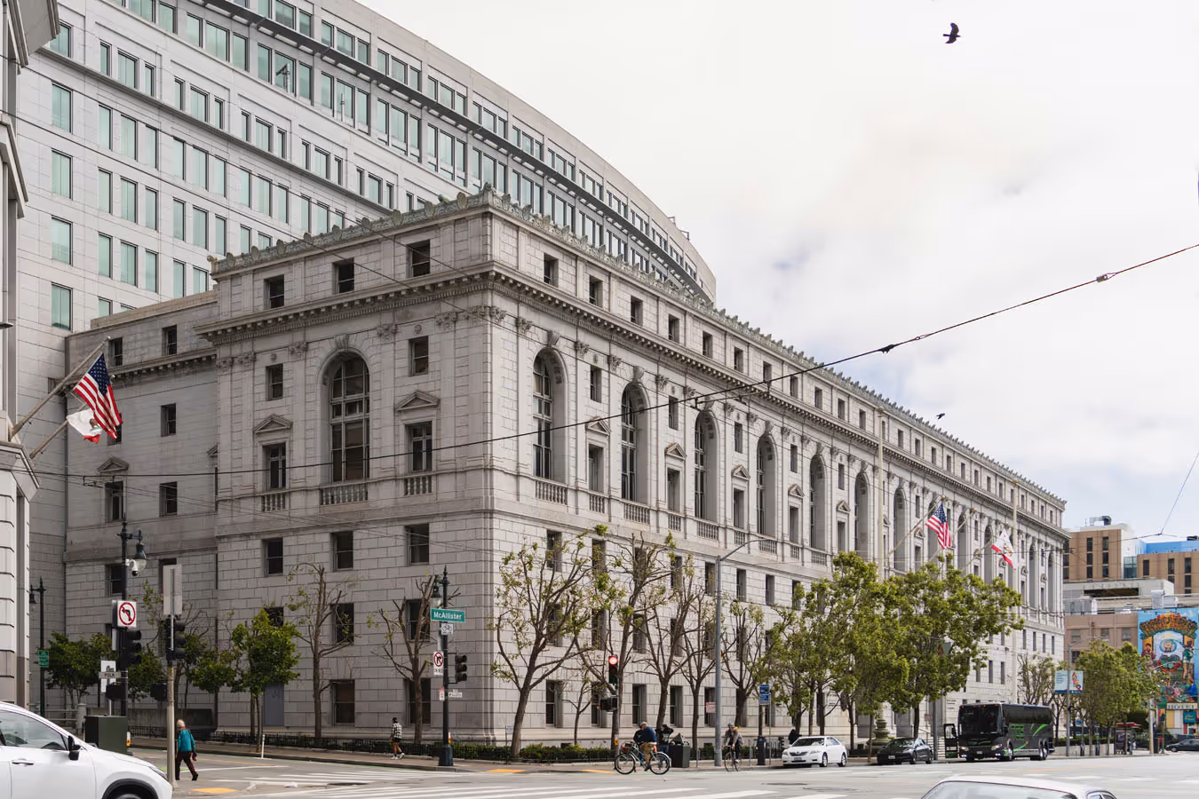 Stone government building with arched windows and American flags at a street intersection with trees and pedestrians.