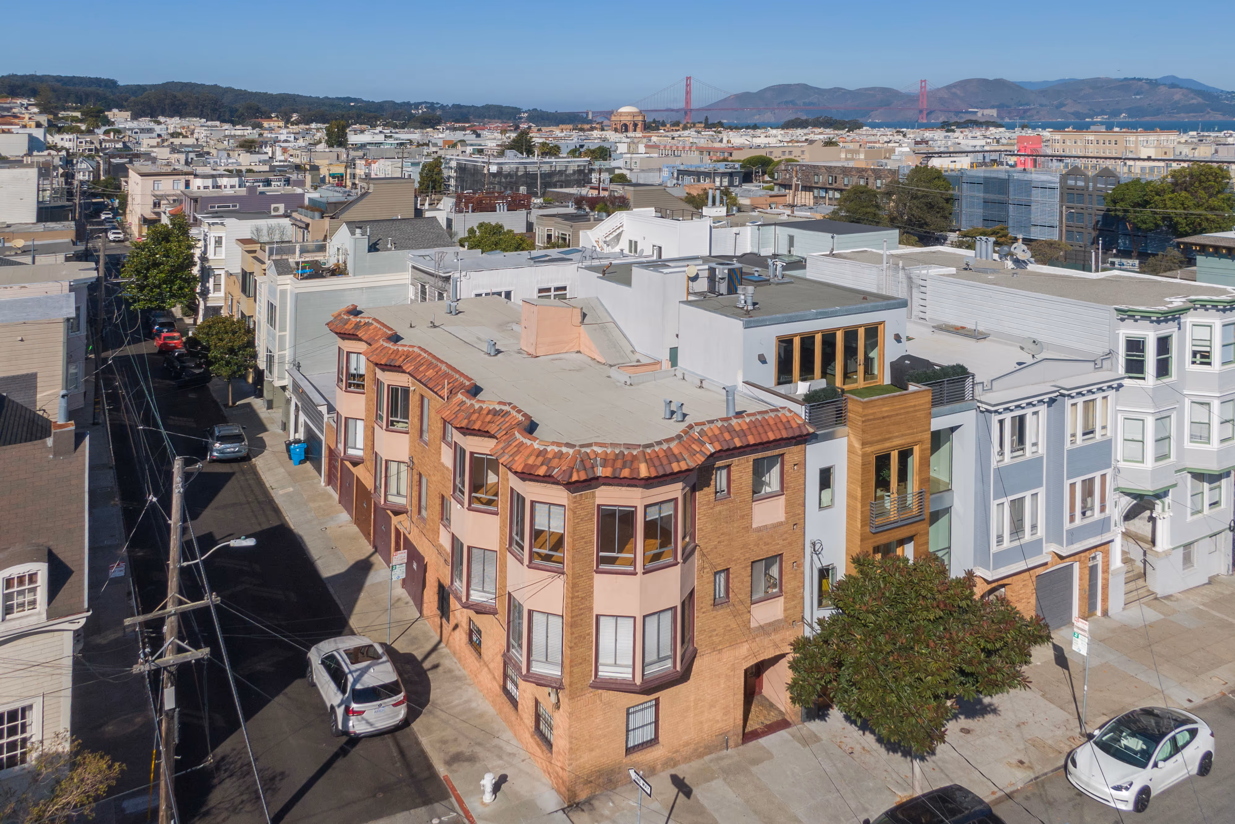Aerial view of a residential building with distinct bay windows and terracotta roof tiles on a sunny day in San Francisco, with the Golden Gate Bridge and Palace of Fine Arts in the background.