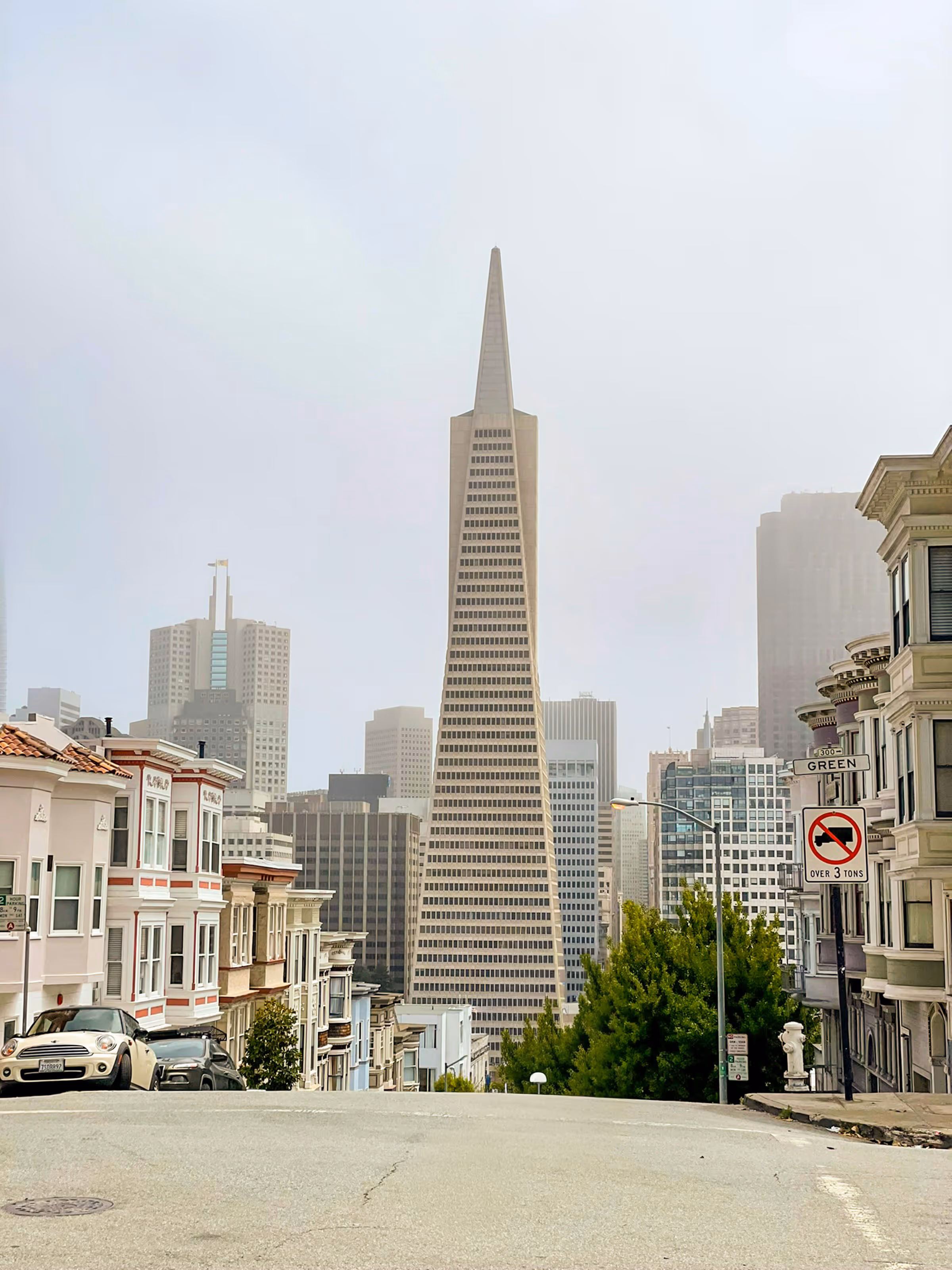 View of the Transamerica Pyramid building in San Francisco from a nearby street with parked cars and residential buildings on both sides.