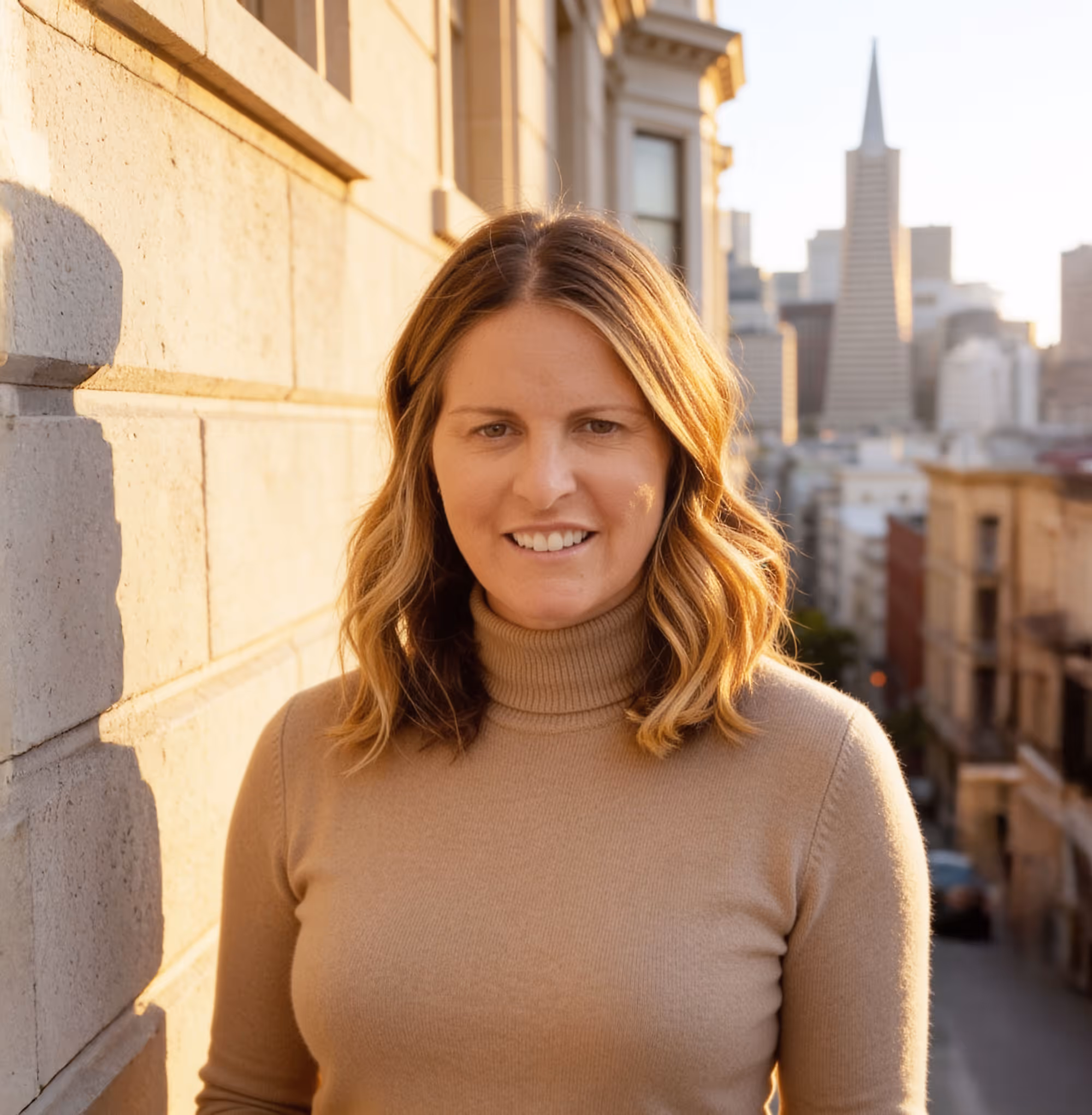 Allison Chapleau with shoulder-length wavy hair wearing a beige turtleneck, standing outdoors in front of a stone wall with a city skyline in the background.
