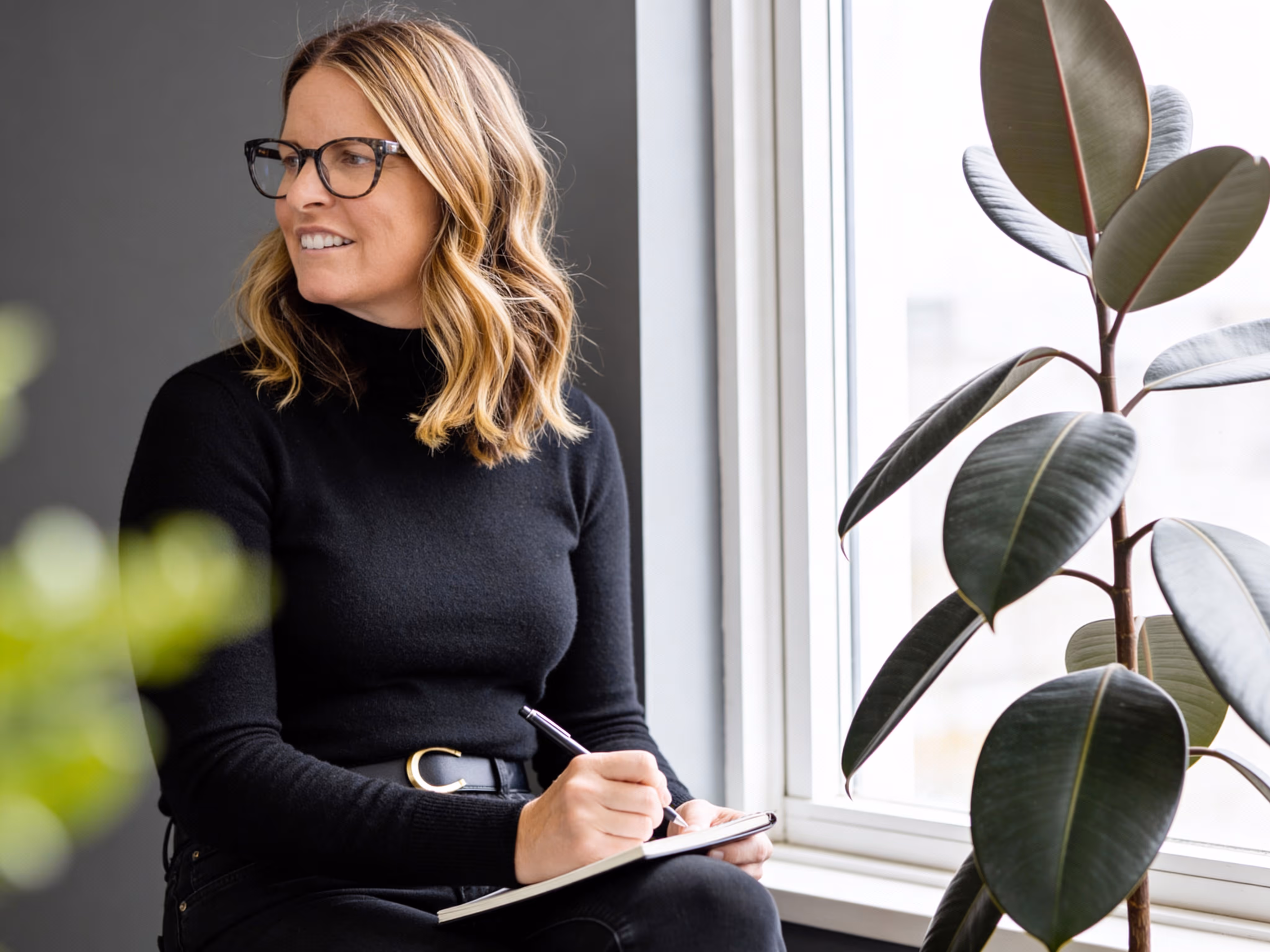 Allison Chapleau wearing black glasses and a black sweater sitting by a window, writing in a notebook, with a large leafy plant nearby.