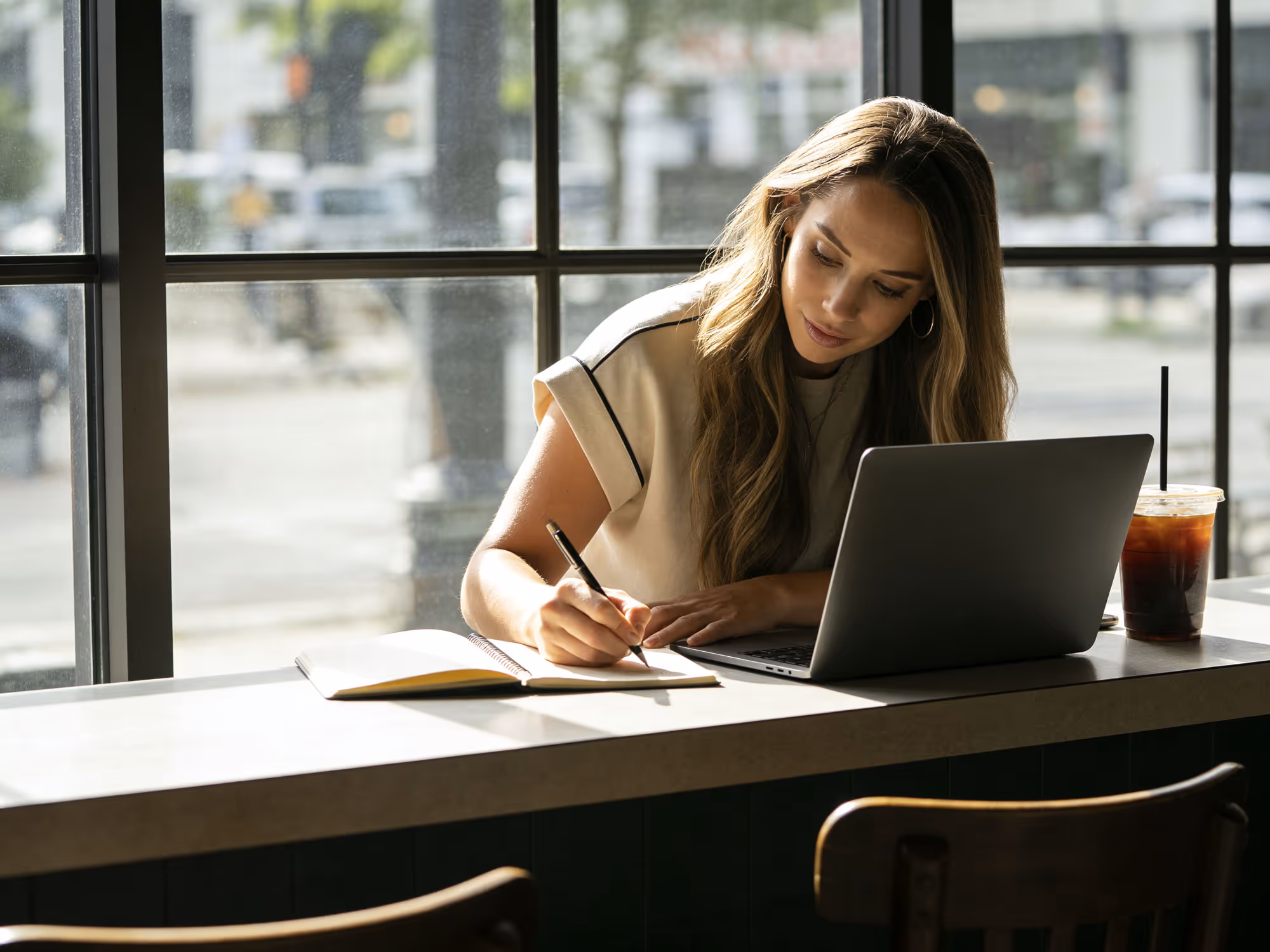 Natalie Meyers writing in a notebook while sitting at a counter with a laptop and iced coffee near a large window.
