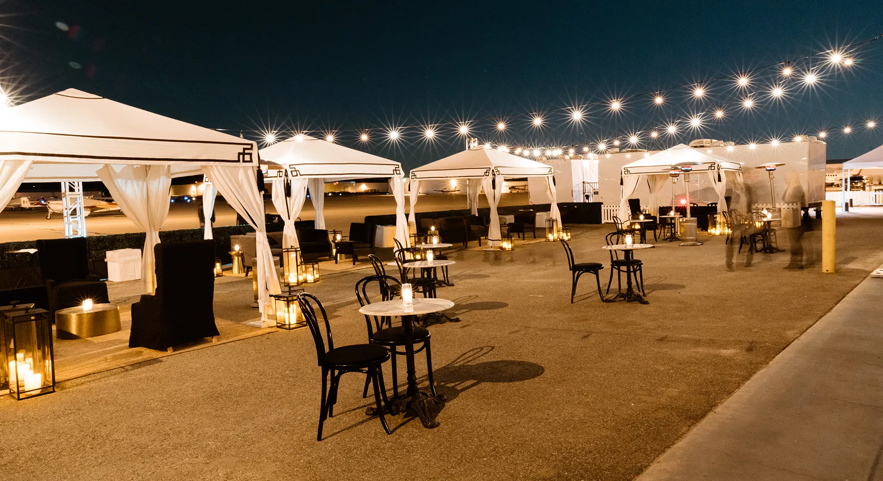 Outdoor cocktail tables and white lounge canopies beneath string lights