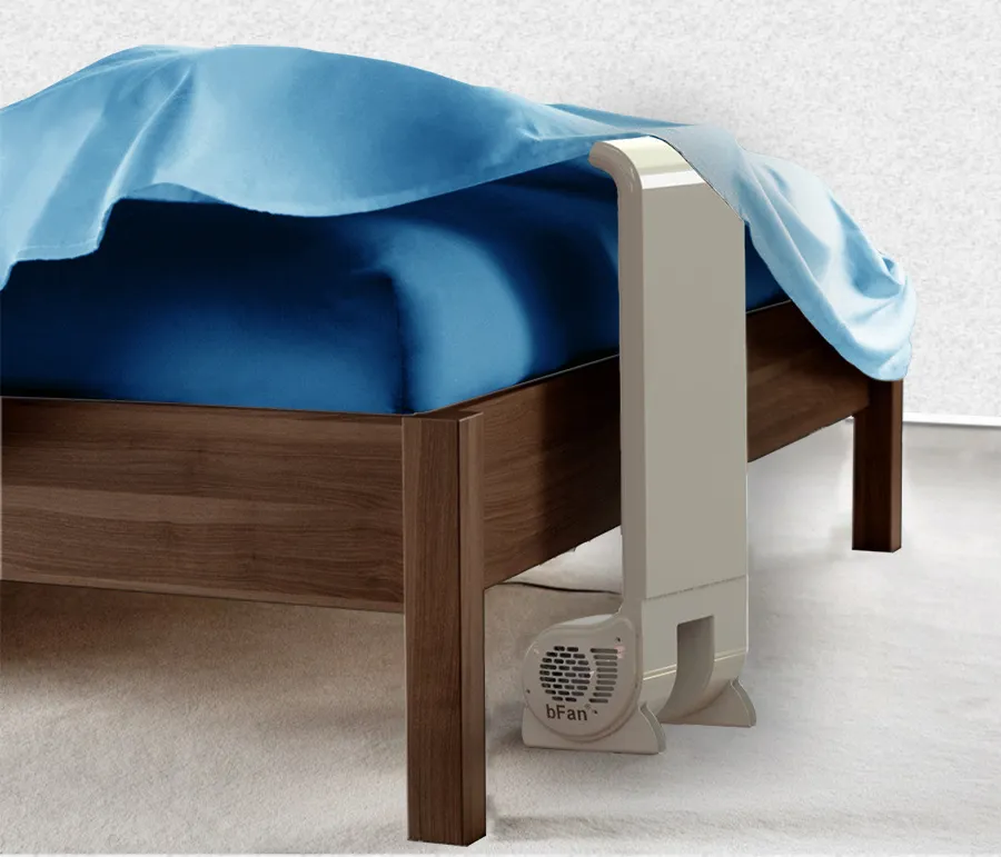 Modern white bFan bed fan placed under a wooden bed frame with blue bedding in a carpeted room.