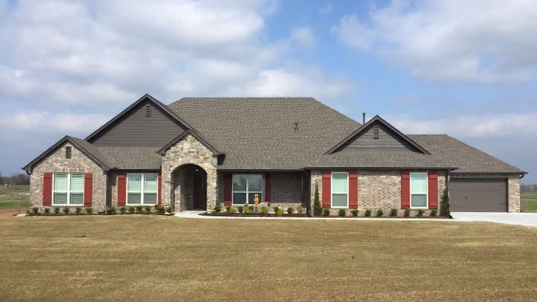 Single-story brick house with brown roof, red shutters, arched stone entryway, and attached garage on a large lawn under a partly cloudy sky.