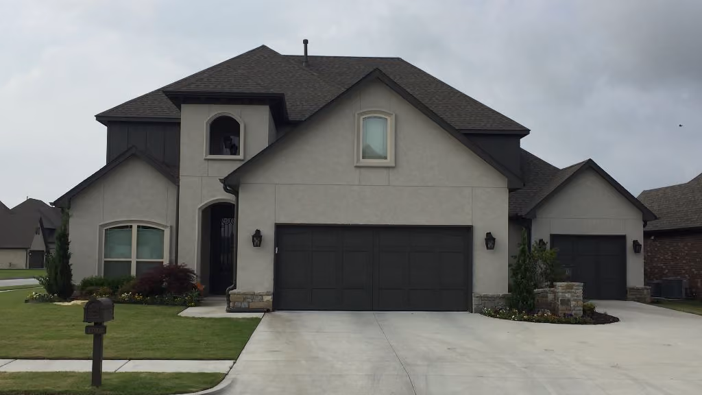 Modern two-story house with gray exterior, dark roof, and two black garage doors on a concrete driveway under a cloudy sky.