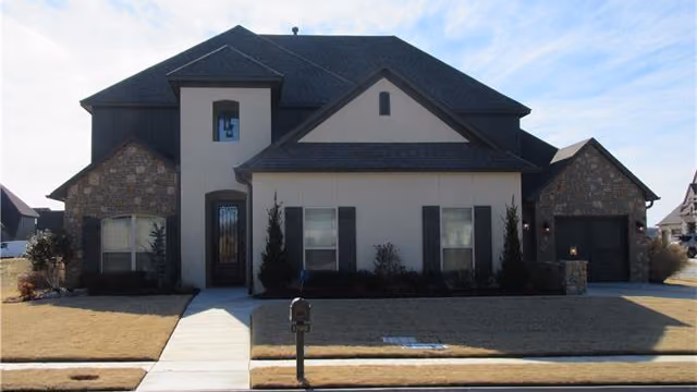 Modern single-family house with a combination of stone and beige stucco exterior, dark roof, and a walkway leading to the front door.