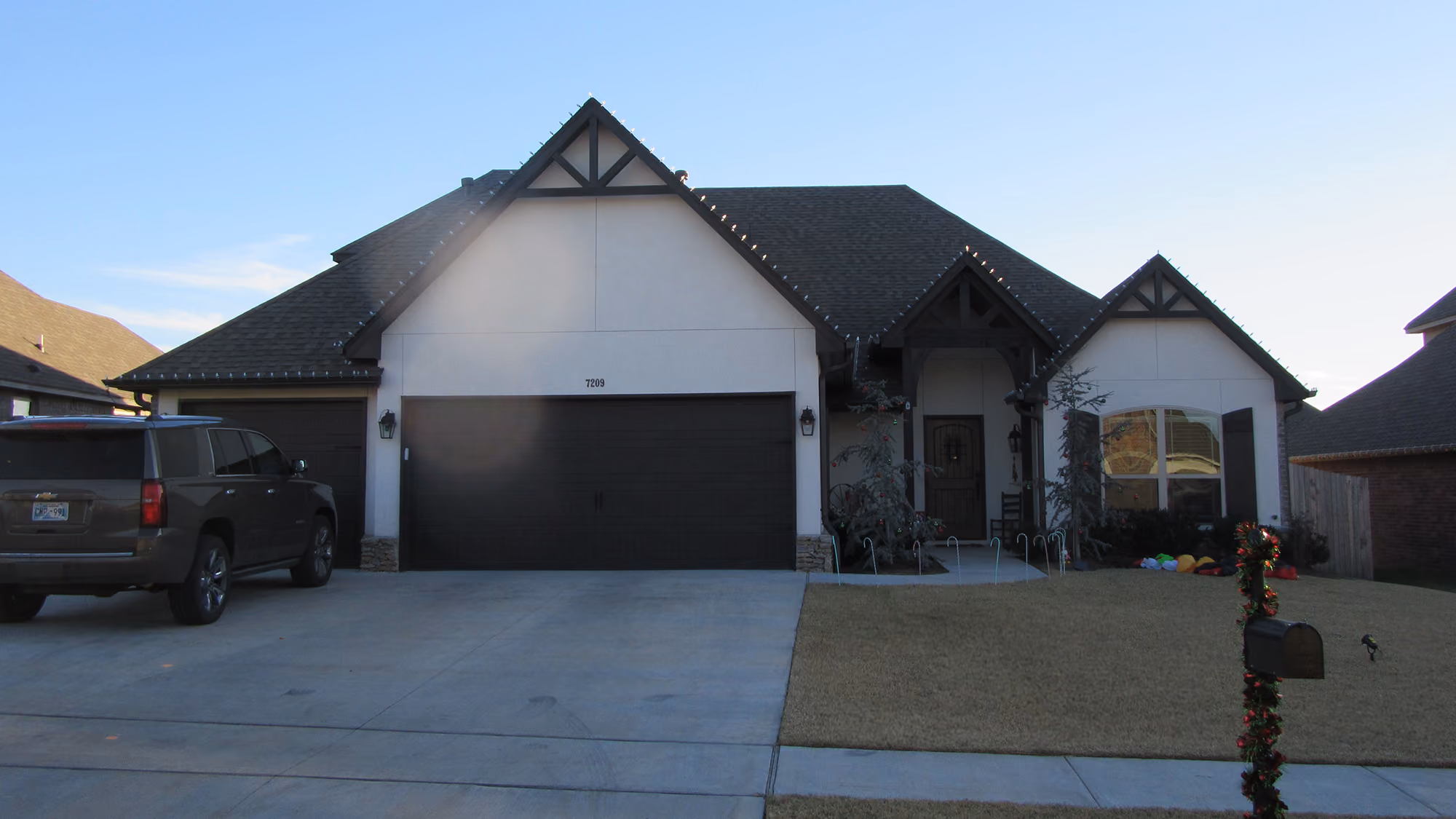 Single-story house with three gabled roof sections, double garage, Christmas decorations on mailbox and shrubs, and an SUV parked on the driveway.