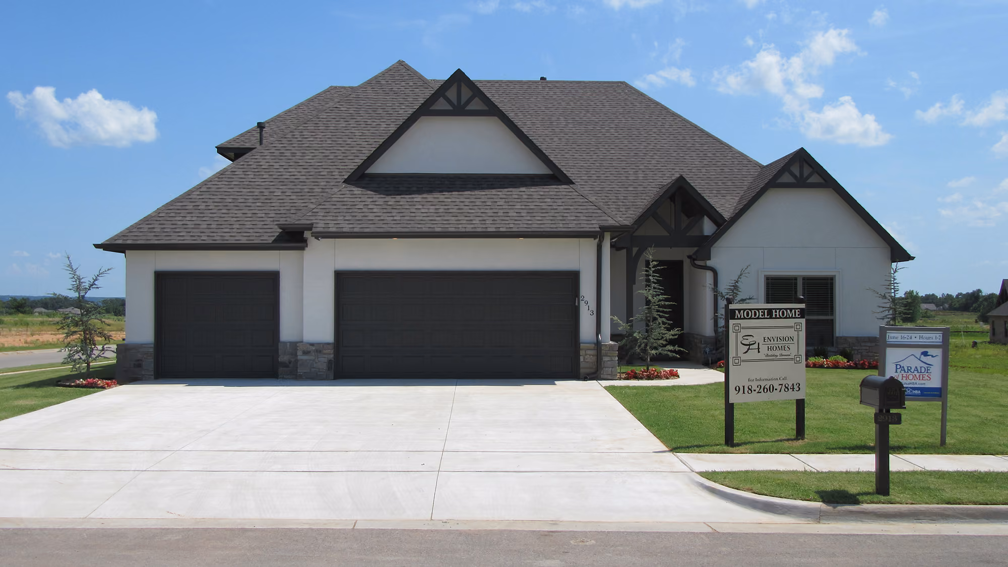 Single-story model home with gray roof, two dark garage doors, stone accents, and a green lawn with signage in front.