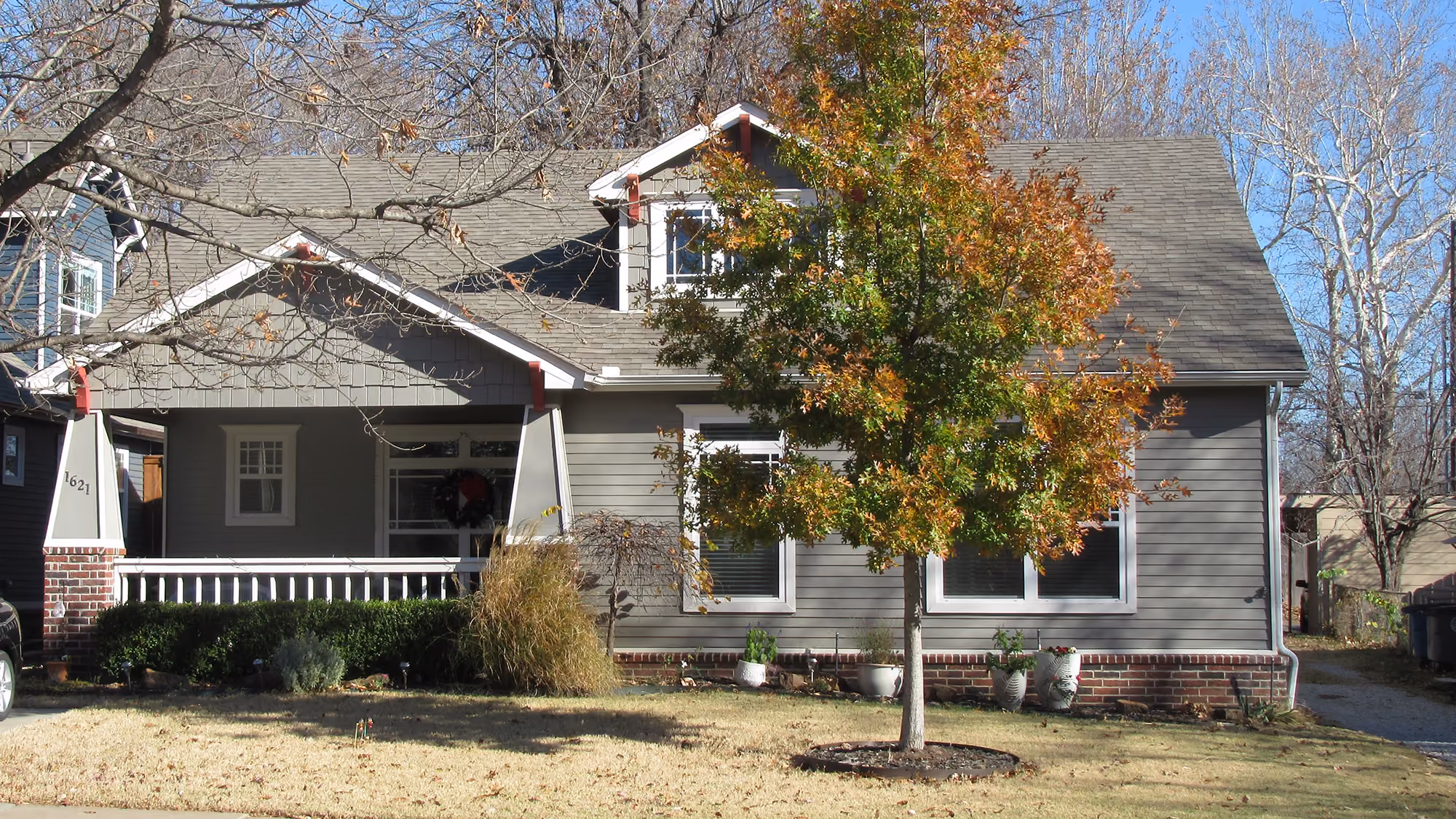 Gray single-story house with a front porch, a tree with autumn leaves, and a lawn in the foreground.