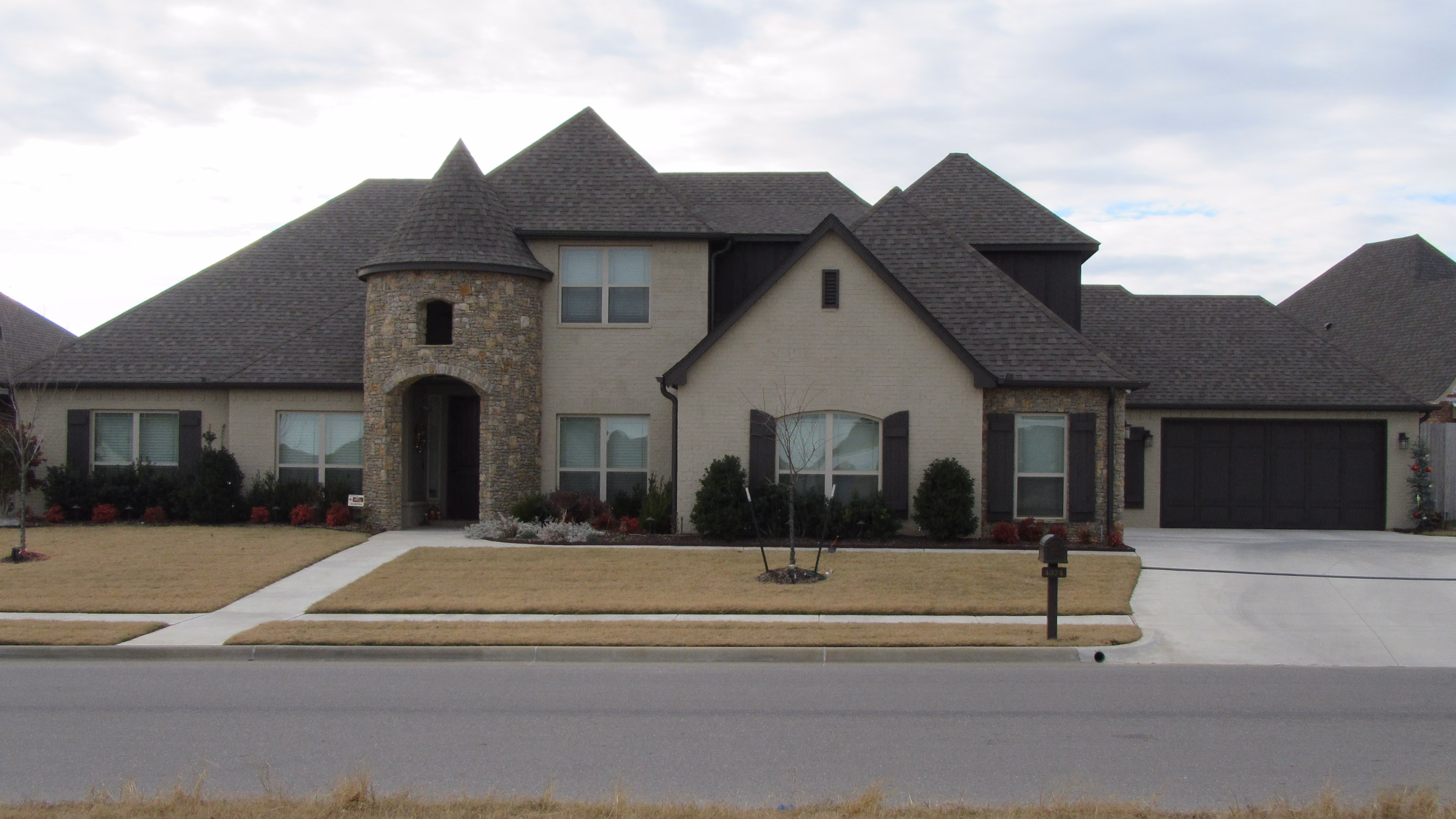 Large beige brick house with dark gray roof, turret entrance, front yard with dry grass, and driveway leading to a two-car garage.