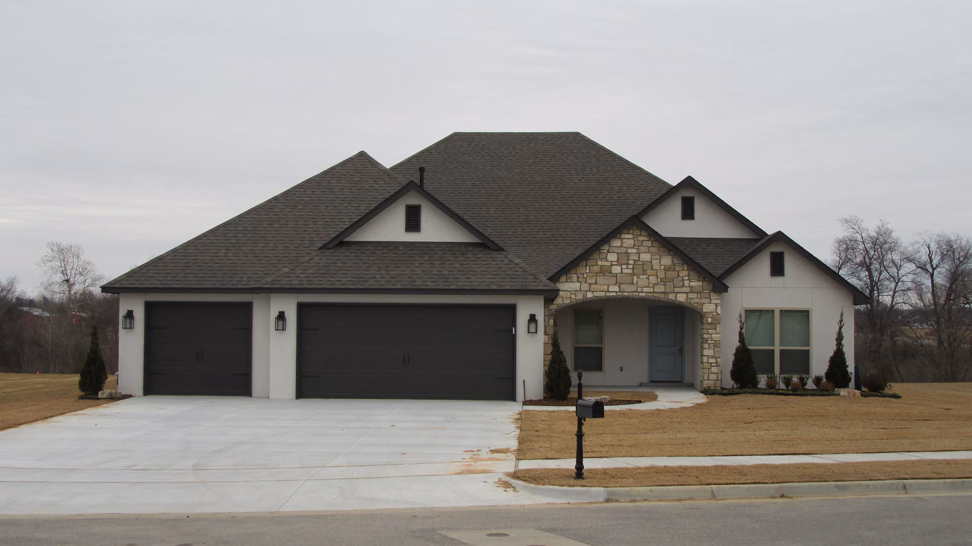 Single-story modern house with three dark garage doors, stone accent around front entrance, and a large concrete driveway under an overcast sky.