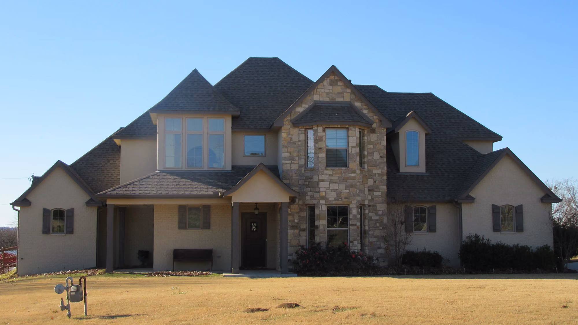 Two-story suburban house with beige walls, a stone facade section, multiple windows, and a dark shingled roof under a clear blue sky.