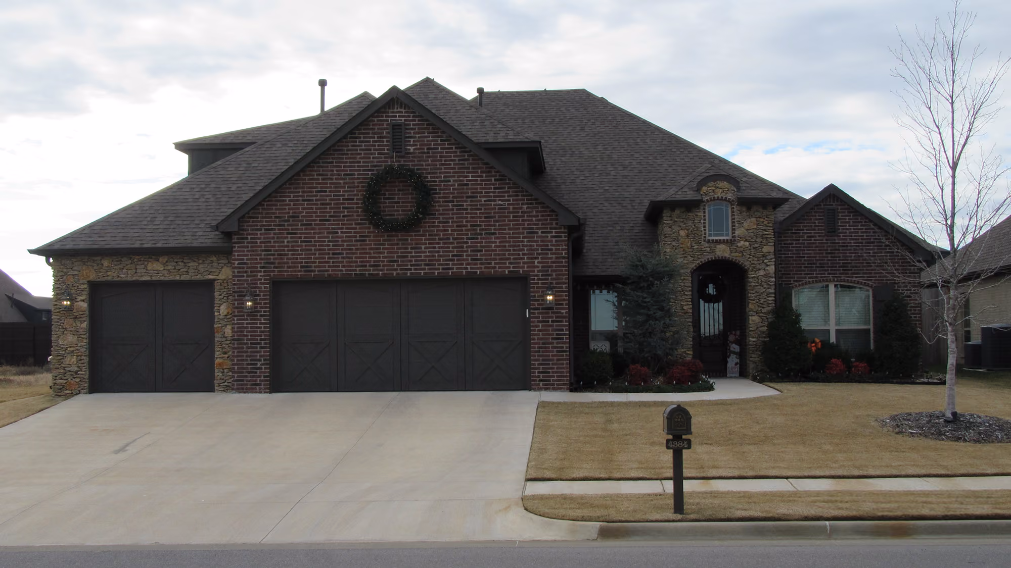 Modern brick and stone single-story house with a three-car garage, a wreath on the garage, a front yard with a tree, and a mailbox.