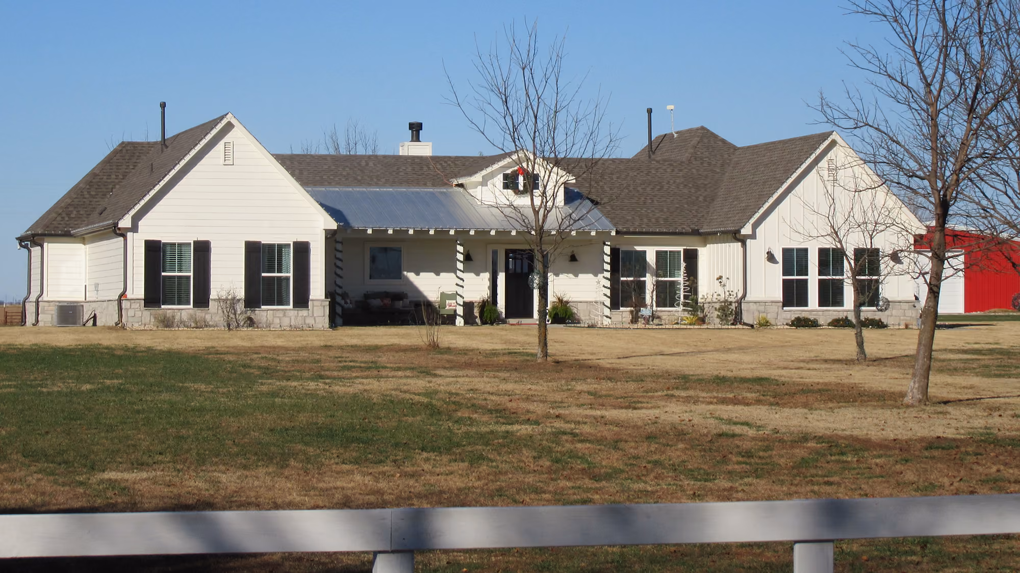 Front view of a single-story white house with dark shutters, a metal porch roof, and a large lawn with leafless trees.