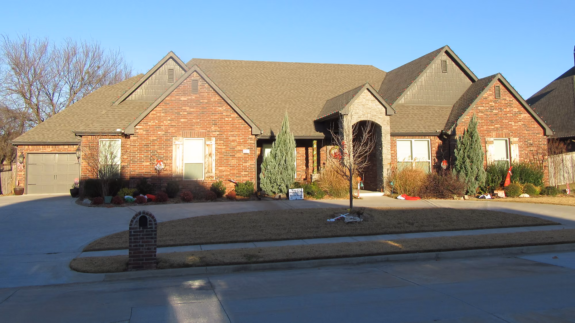 Front view of a large single-story brick house with a two-car garage, a wide driveway, landscaped front yard with bushes and small trees, and clear blue sky.
