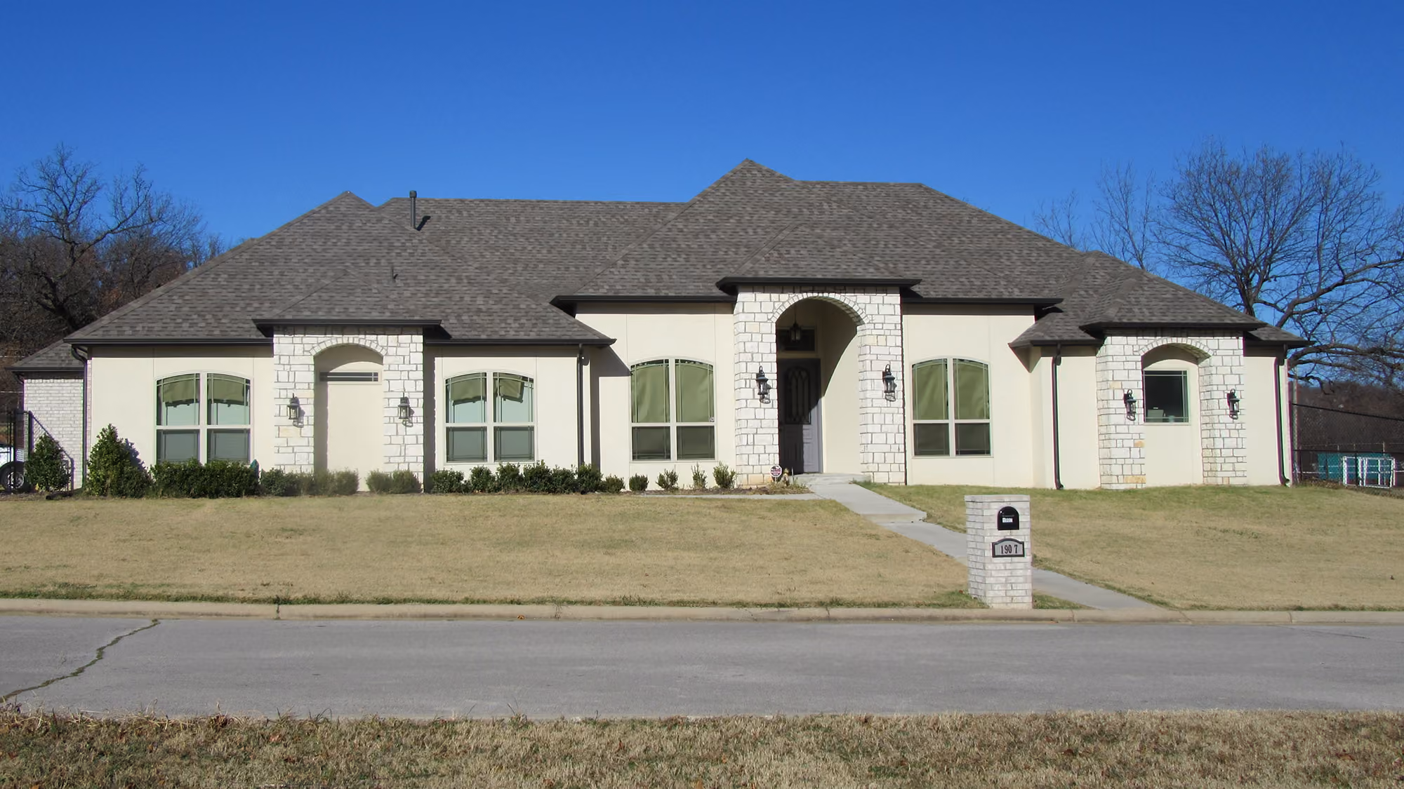 Single-story modern house with stone and stucco facade, large windows with green curtains, and a front lawn with mailbox.