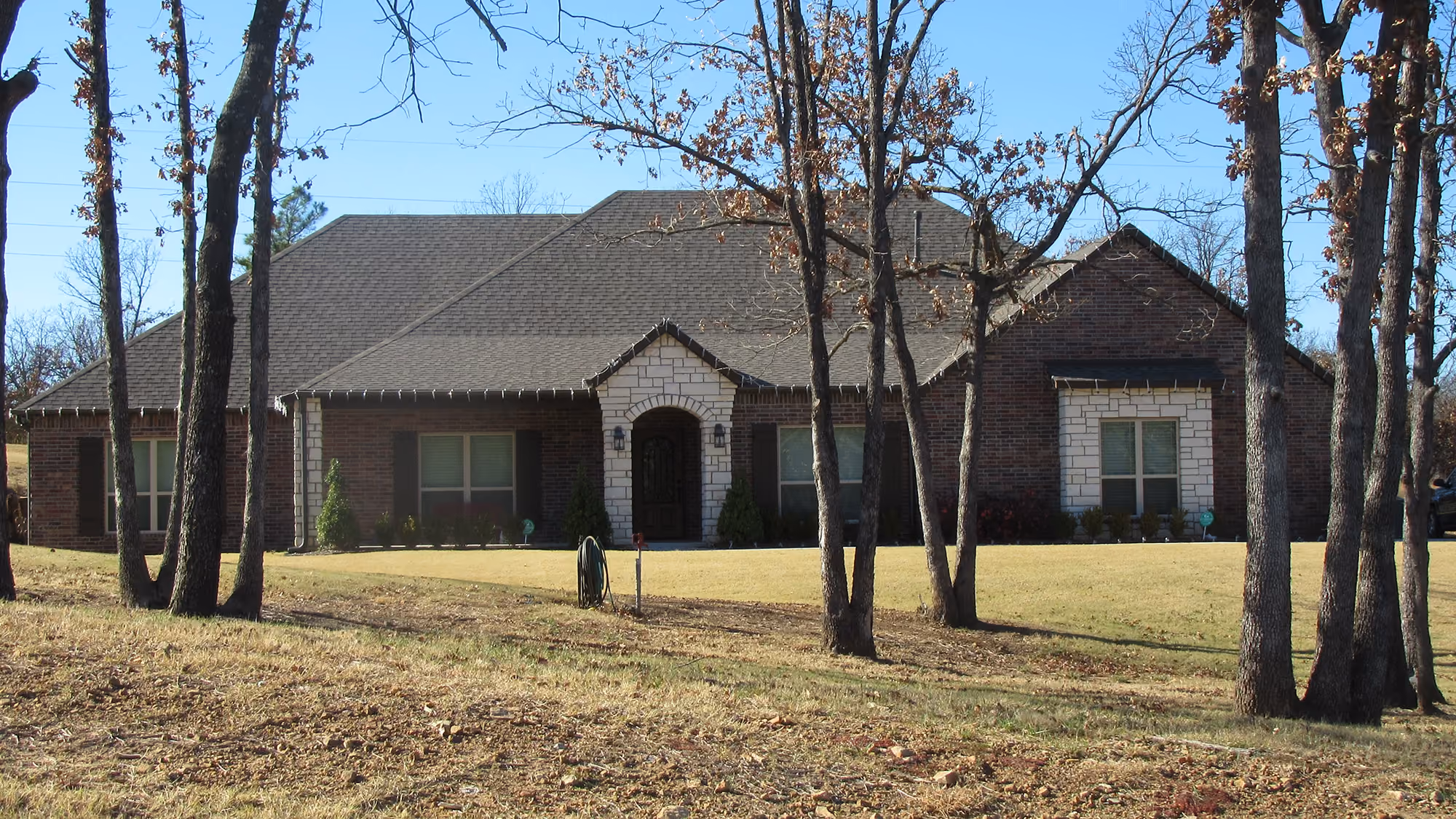 Single-story brick house with stone accents, large front lawn, and leafless trees in front under a clear blue sky.