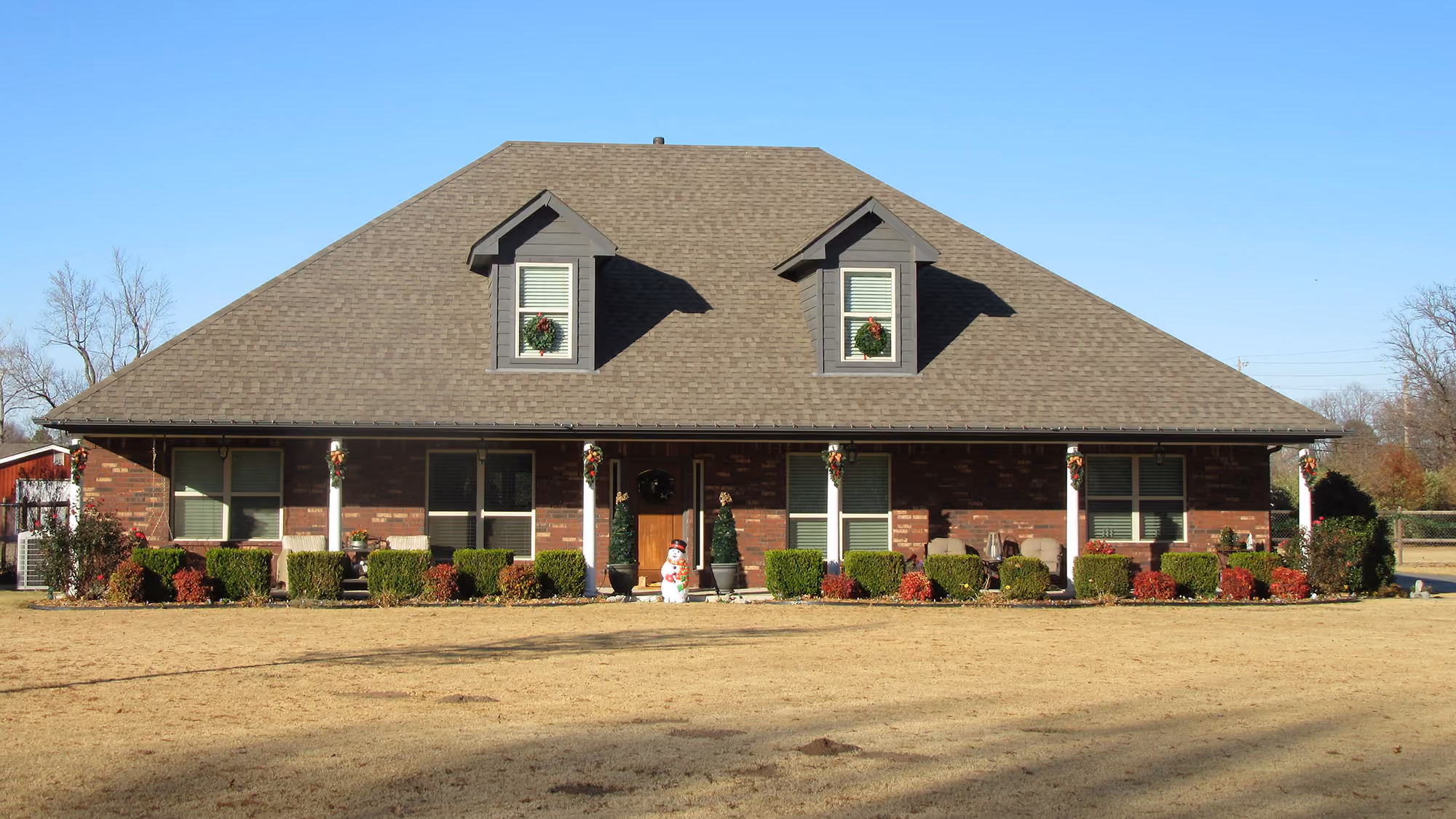 Single-story brick house with a large hipped roof, two dormer windows with wreaths, white columns with holiday decorations, and a snowman figure by the front door.