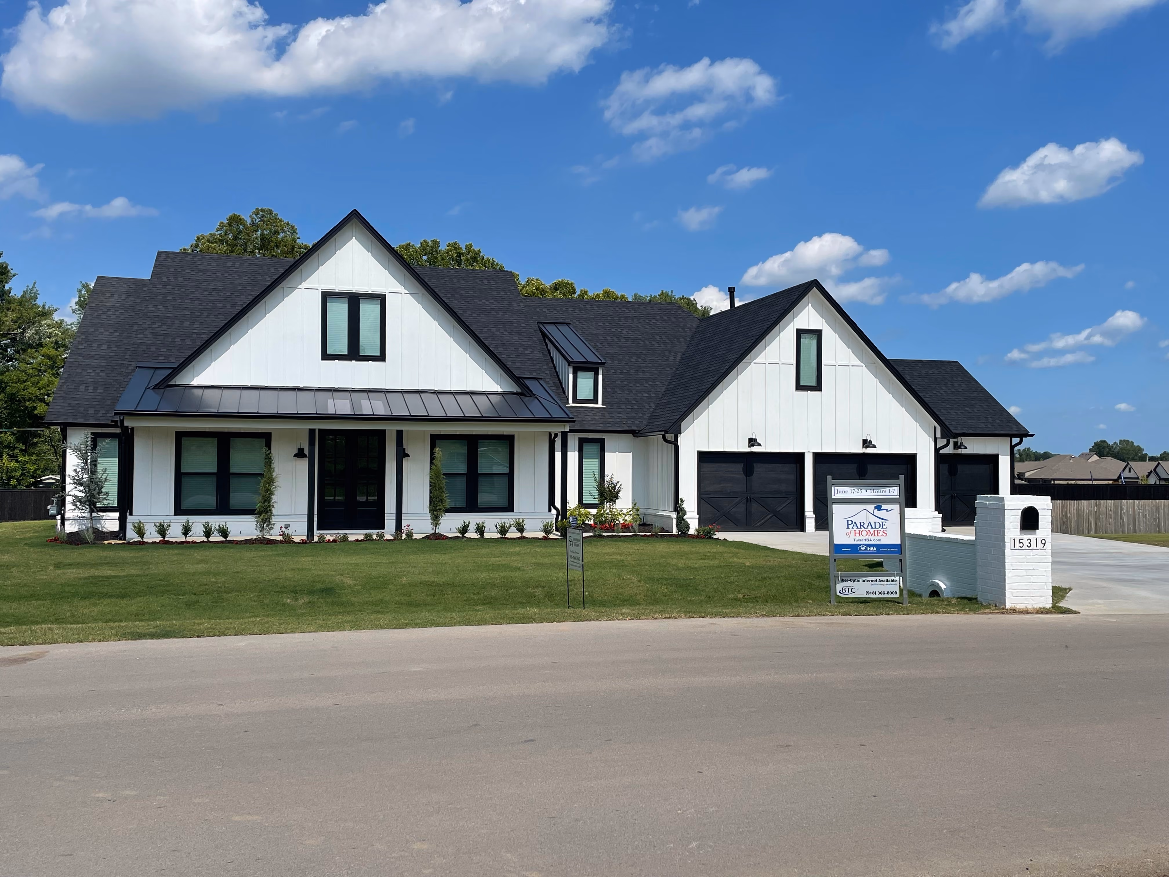 Modern white house with black roof and garage doors, green lawn, and a Parade of Homes sign in the front yard.