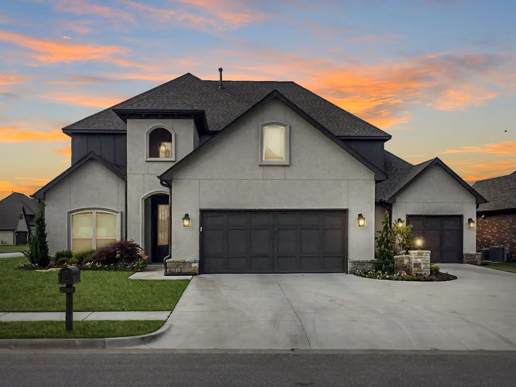 Modern two-story house with gray exterior, dark roof, and two black garage doors under a colorful sunset sky.