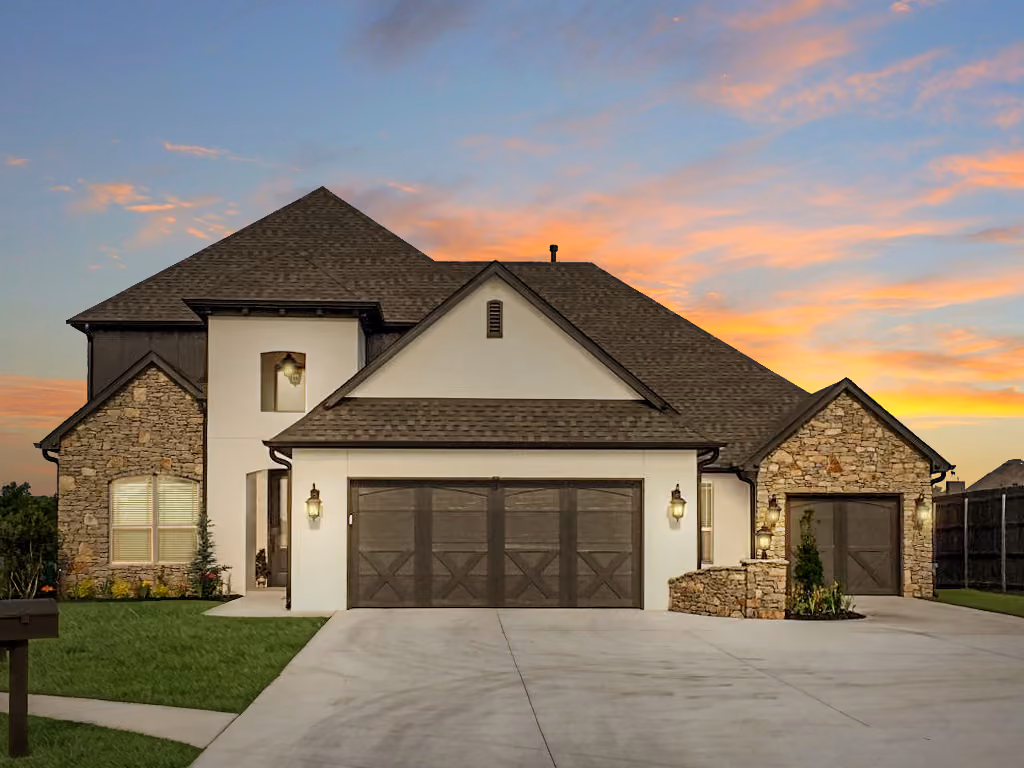 Two-story modern house with stone and white exterior, three-car garage, and a sunset sky background.