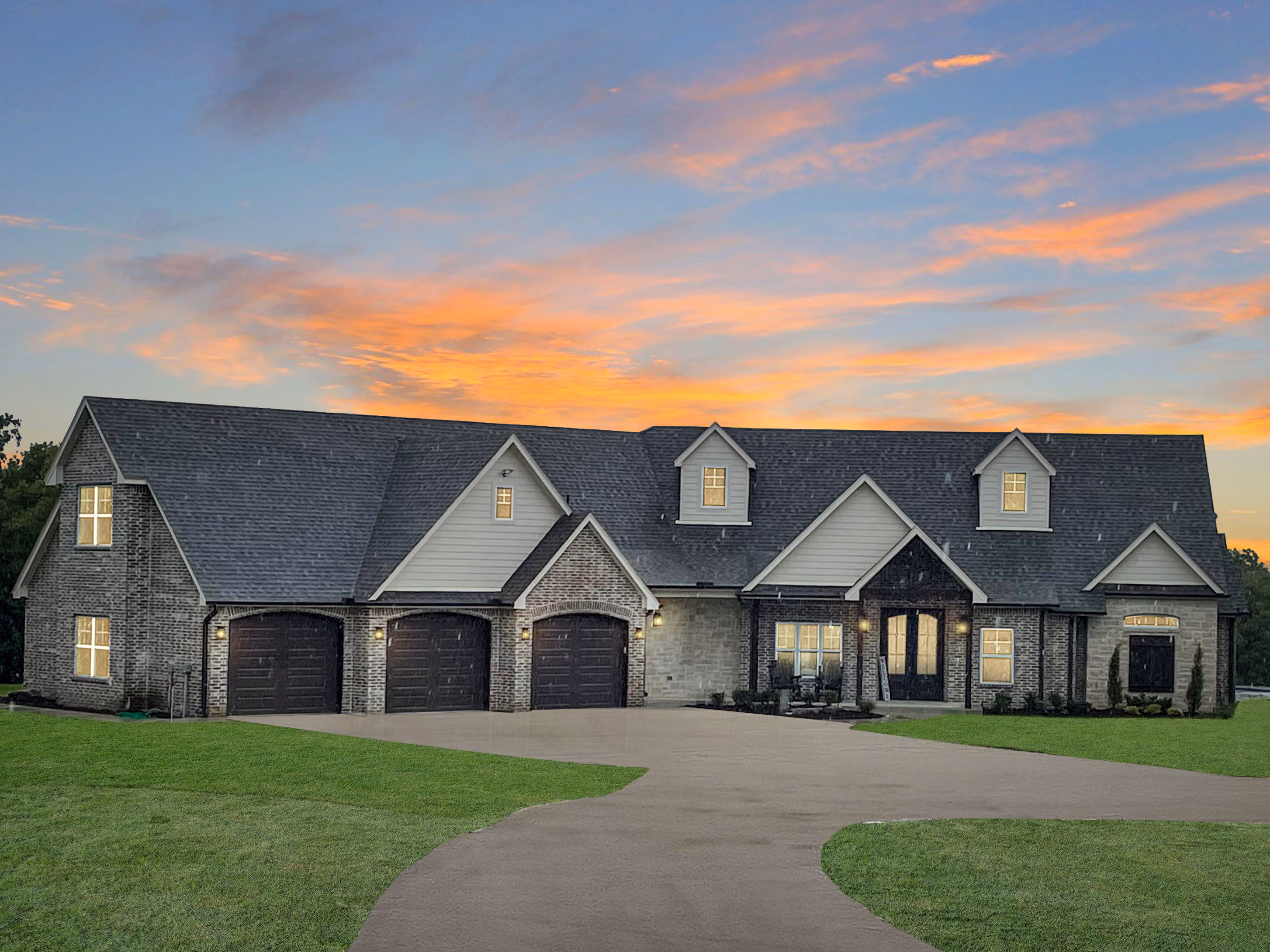 Large modern brick house with three dark garage doors, front porch, and illuminated windows at sunset.