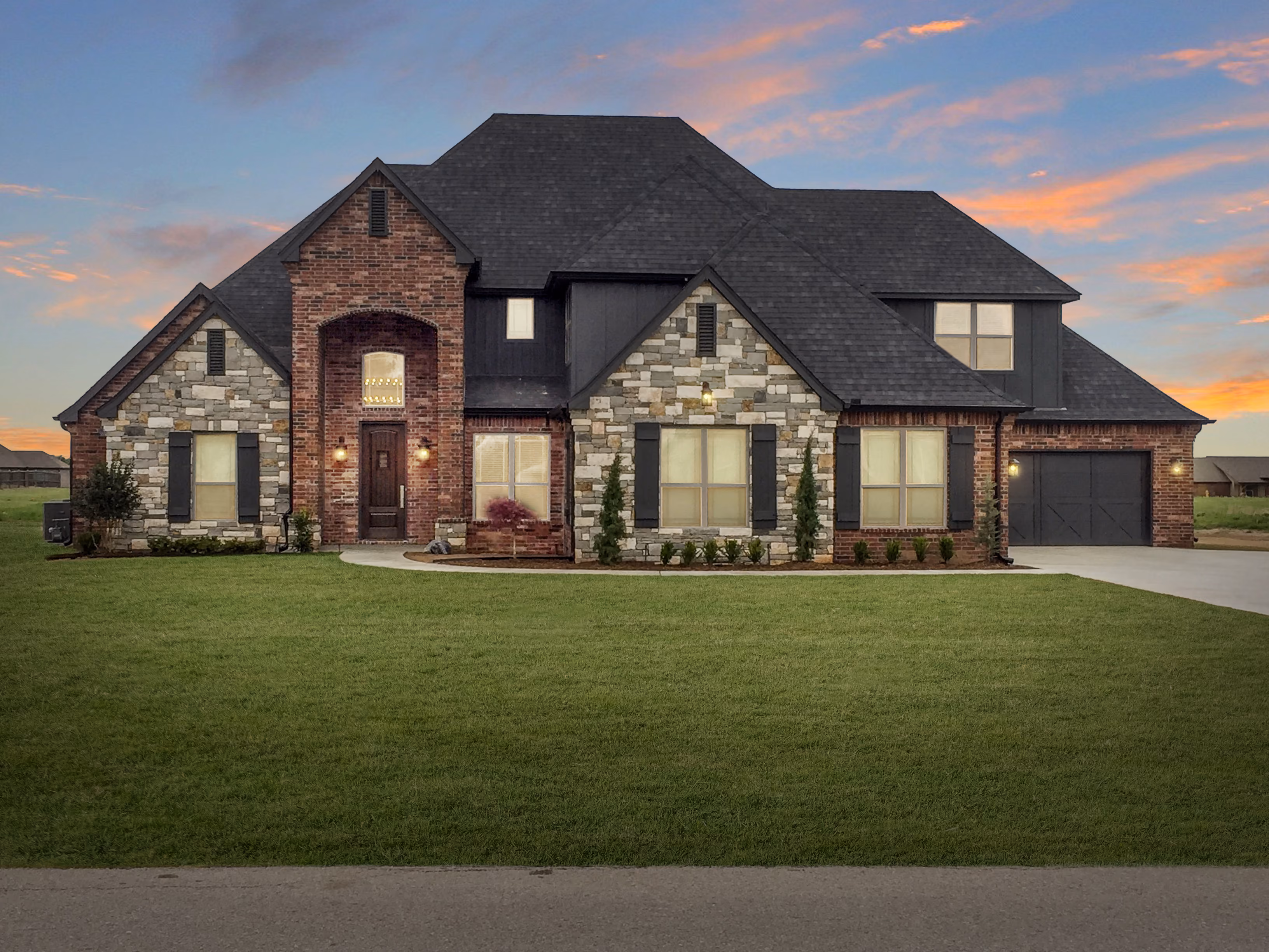 Large modern house with brick and stone facade, black shutters and garage, set against a colorful sunset sky.