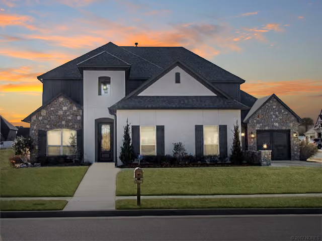 Two-story house with stone and white siding exterior, black roof, front yard with green lawn, and a mailbox at sunset.