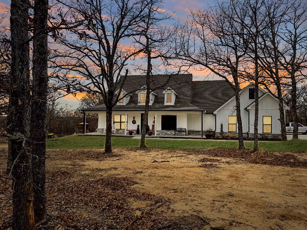 White modern farmhouse with dark roof and stone accents, viewed through leafless trees at sunset.
