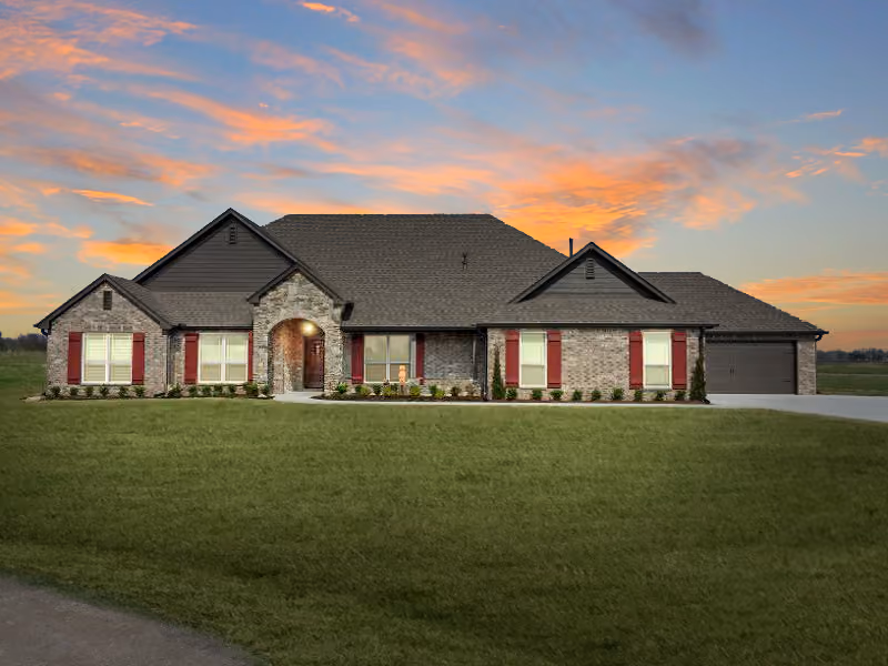 Single-story brick house with brown roof, red shutters, landscaped front yard, and a cloudy sunset sky.