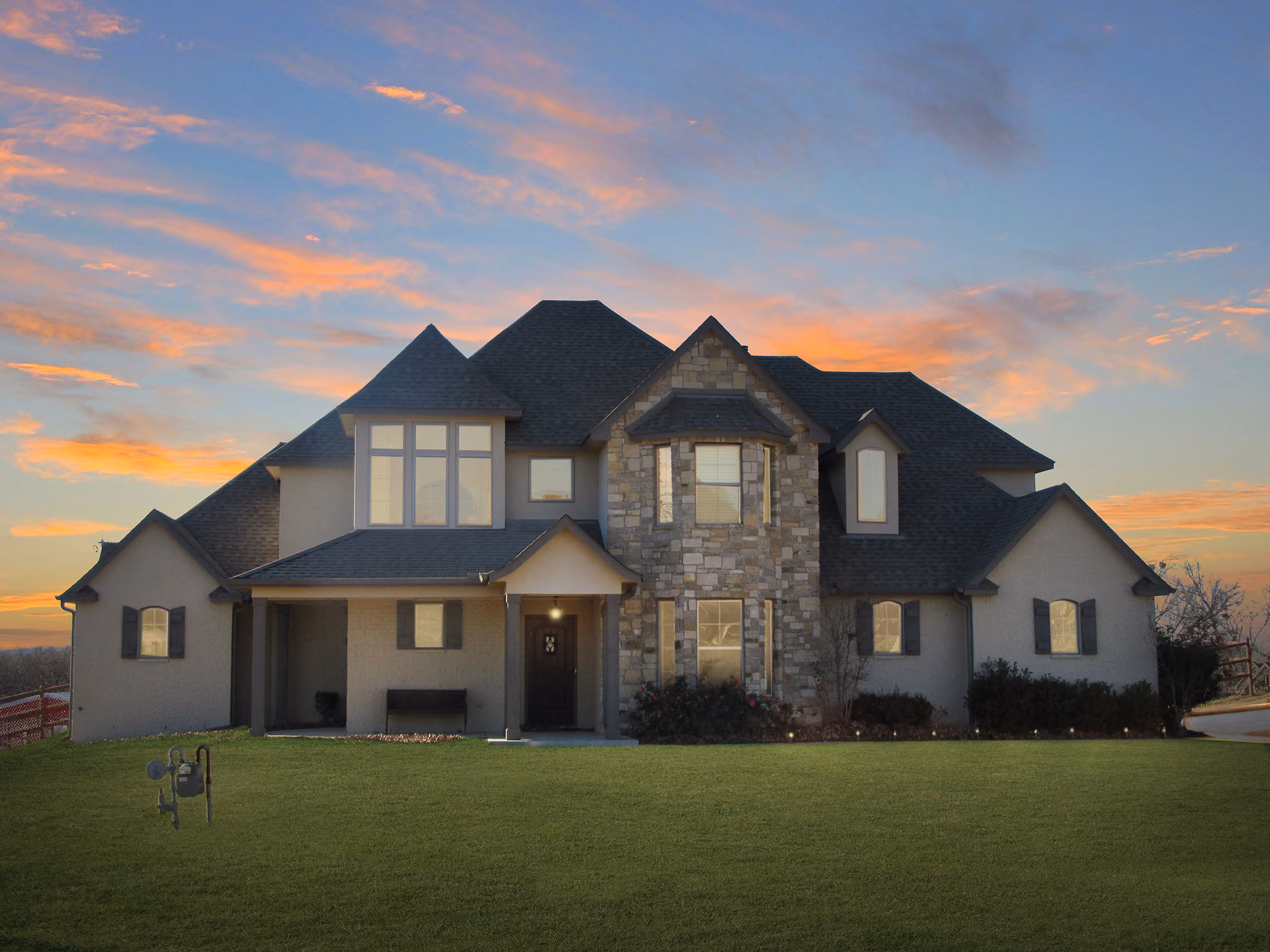 Large two-story house with stone and stucco exterior, multiple gabled roofs, and a green lawn at sunset.
