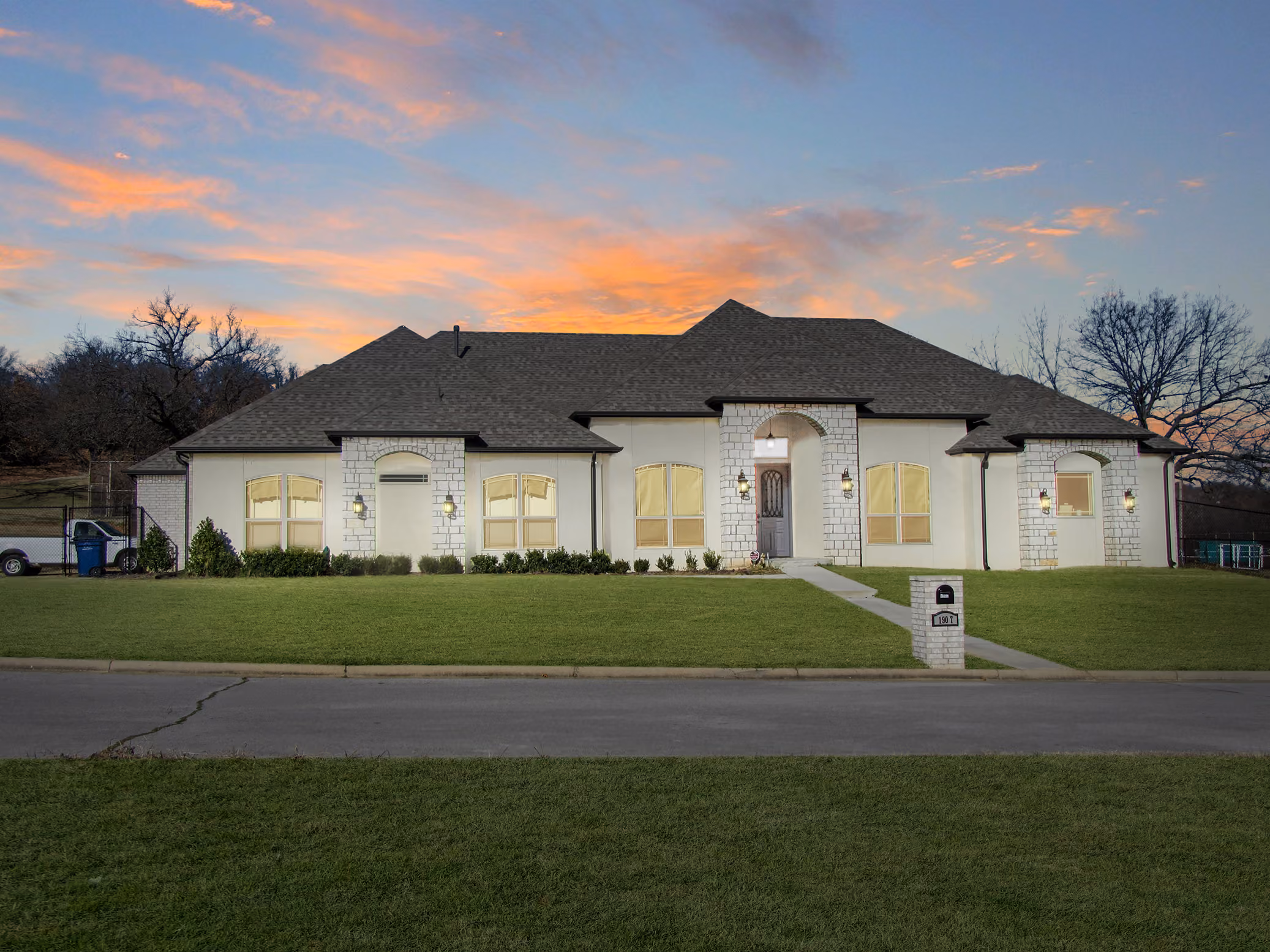 Single-story modern house with a dark roof, stone accents, front lawn, and a mailbox against a sunset sky.