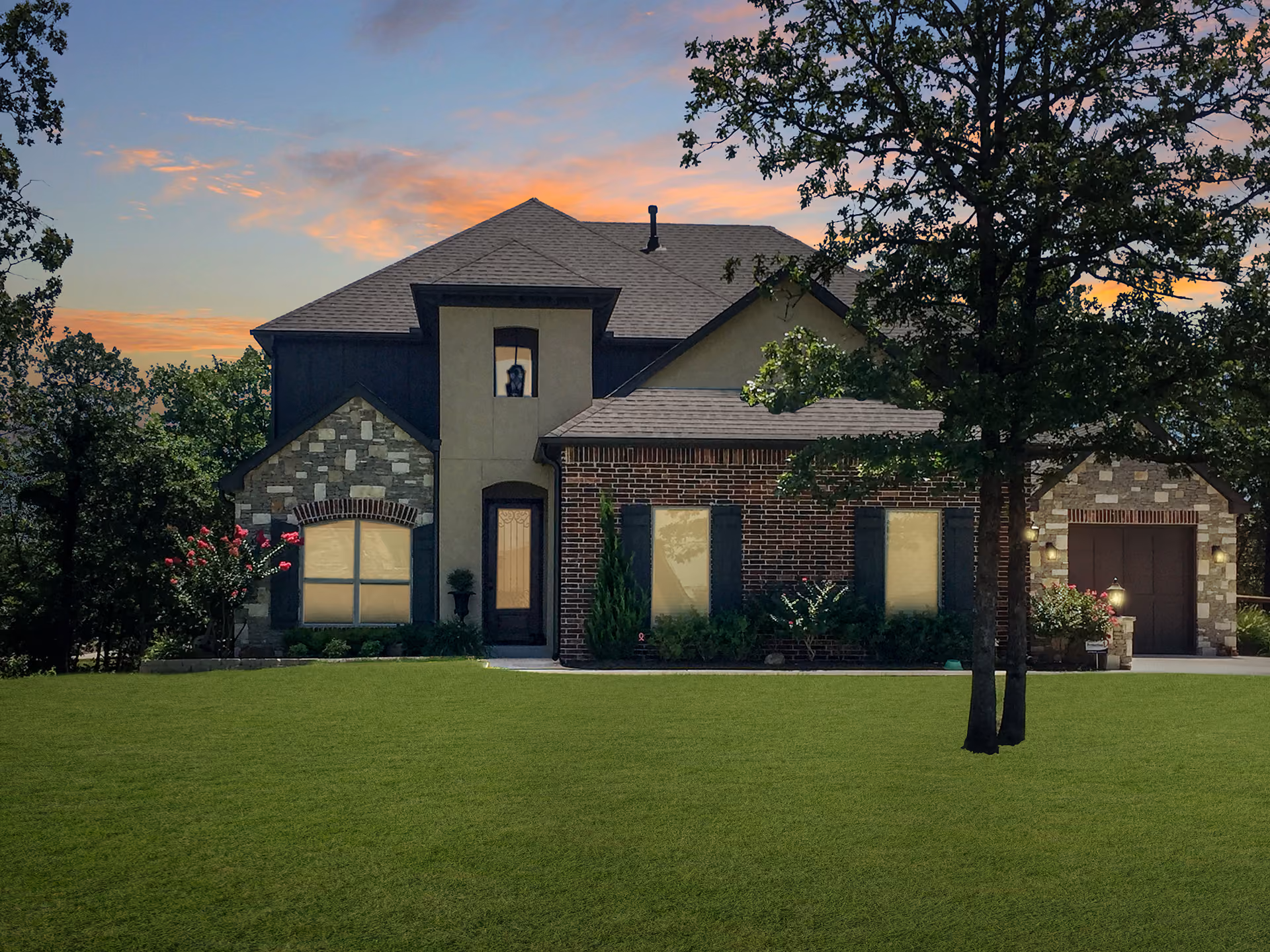 Modern two-story house with brick, stone, and stucco exterior under a sunset sky, surrounded by trees and a green lawn.