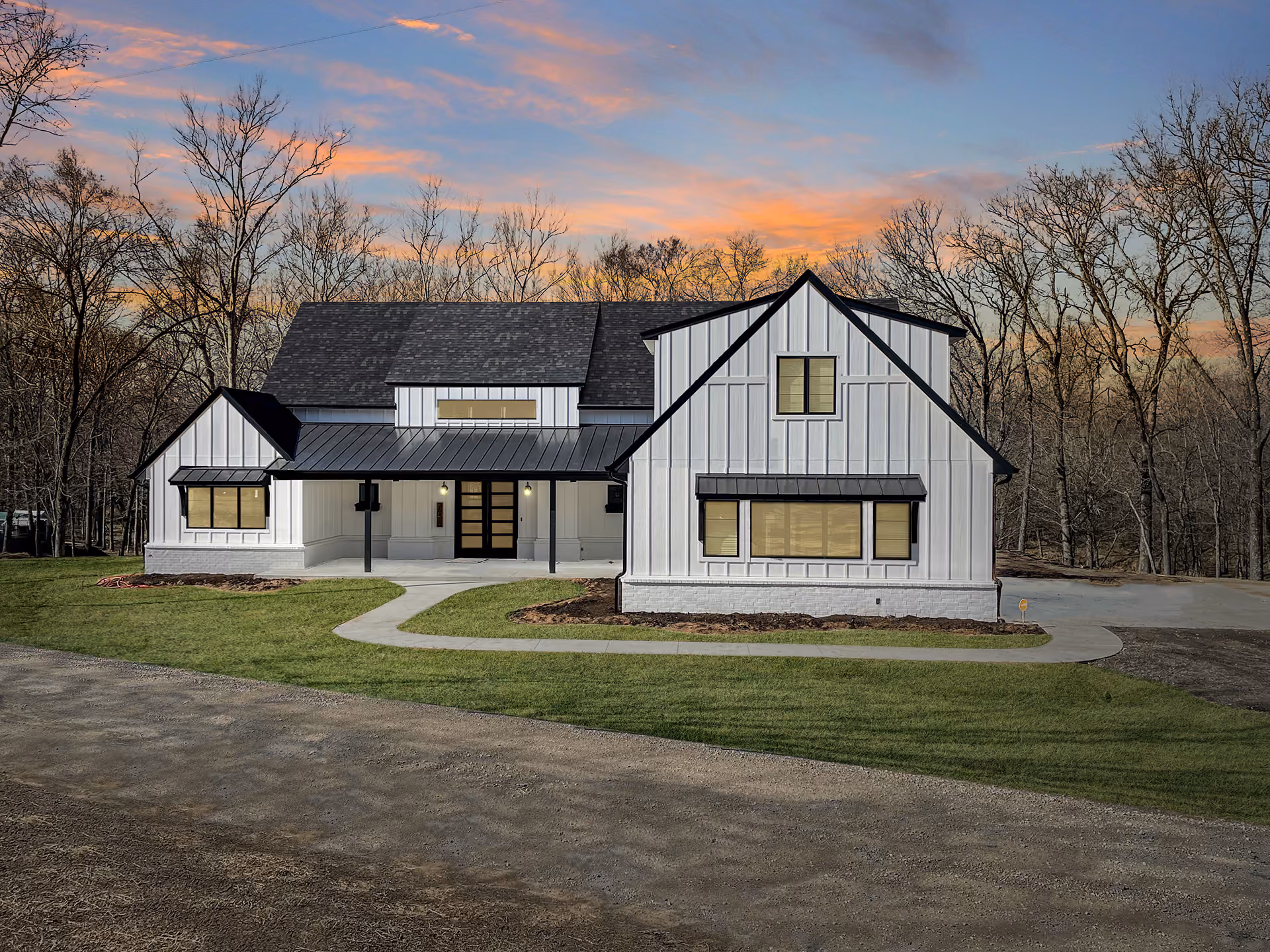 Modern white farmhouse with black roof and window accents, surrounded by bare trees at sunset.