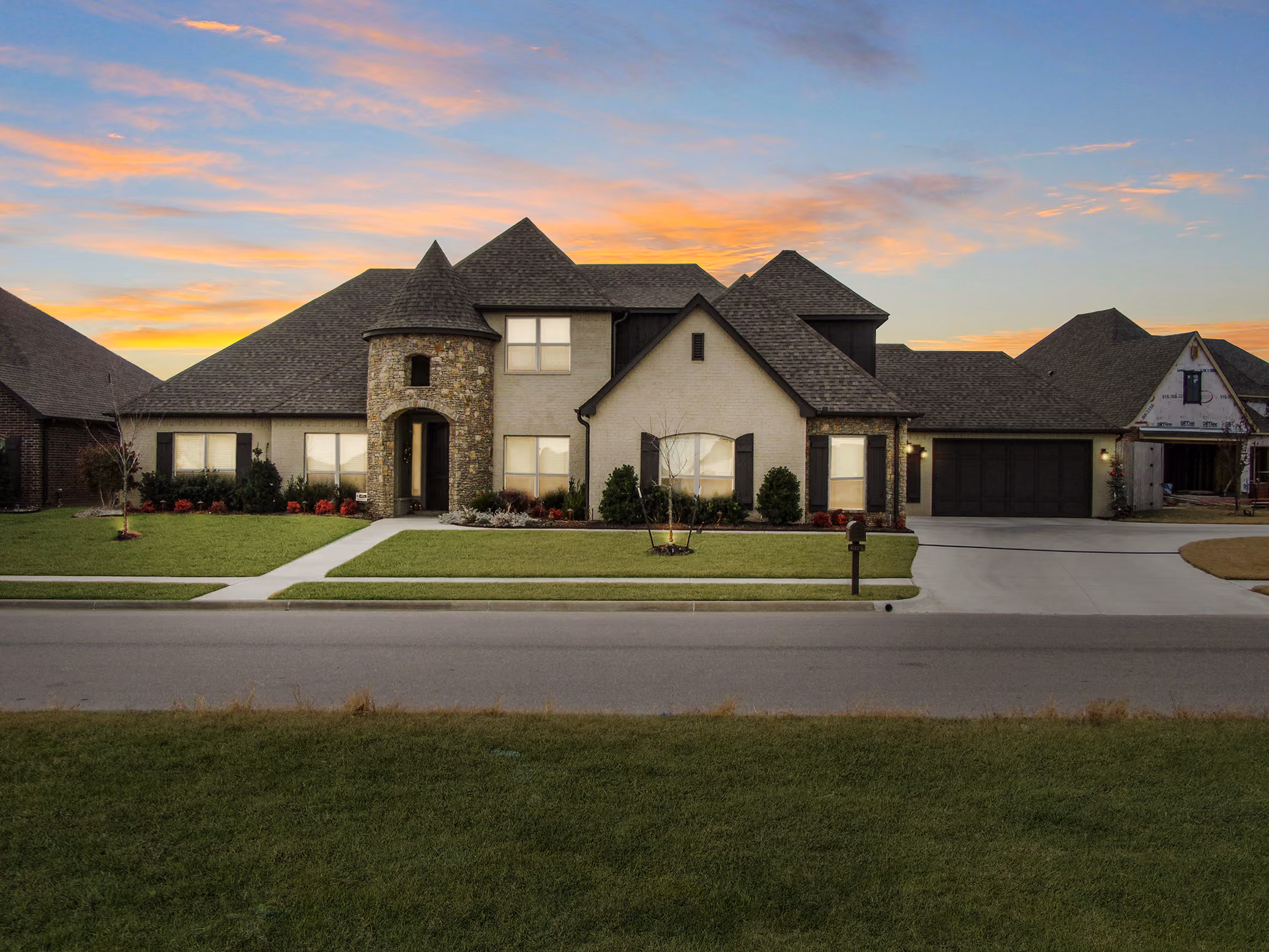 Large suburban house with stone and beige exterior, turret entrance, black shutters, and attached garage at sunset.